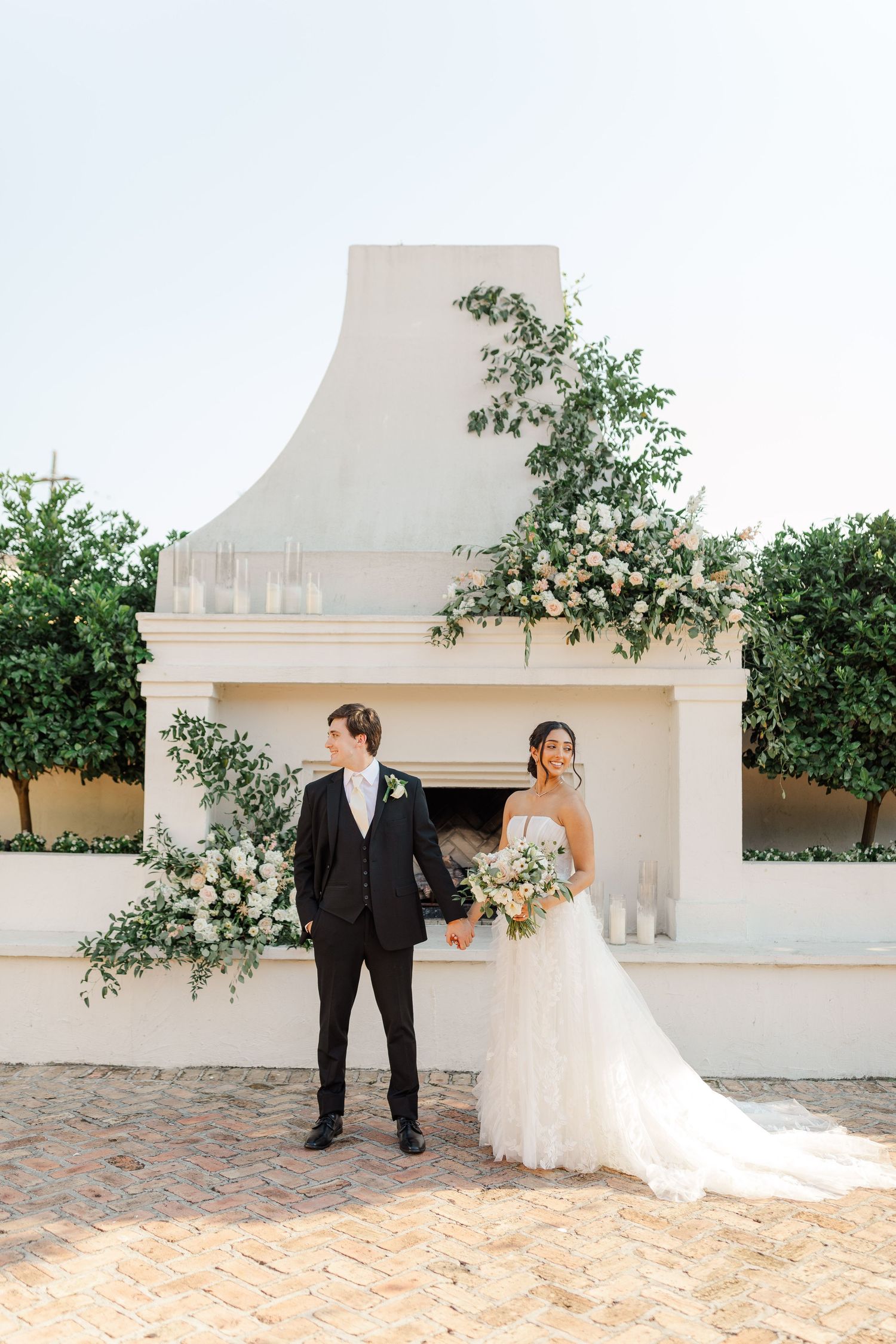 Wedding couple stands before elegant white fireplace adorned with climbing greenery and floral arrangements.