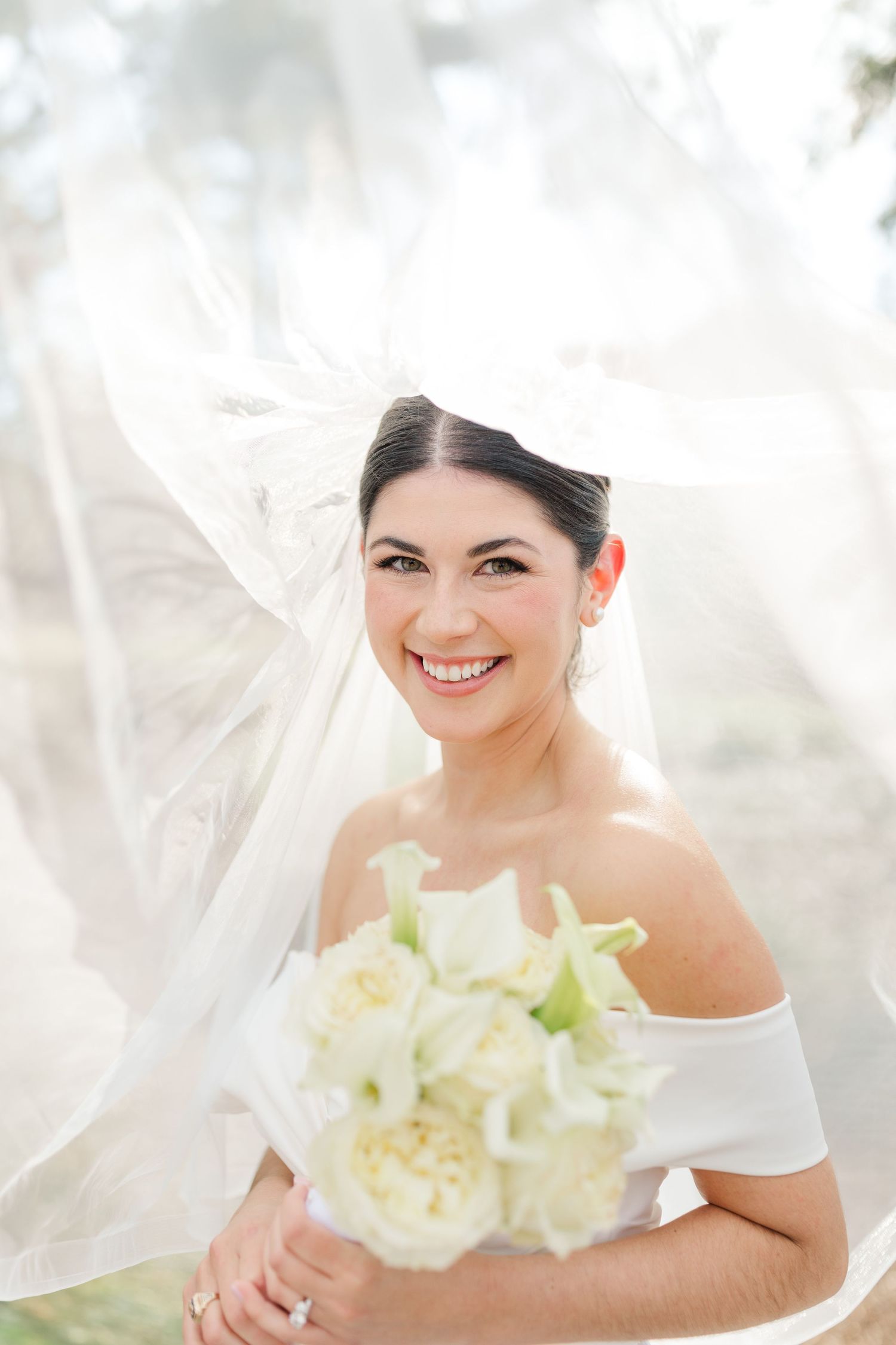 Radiant bride in off-shoulder dress holds elegant white calla lily bouquet while veil flows in soft light.