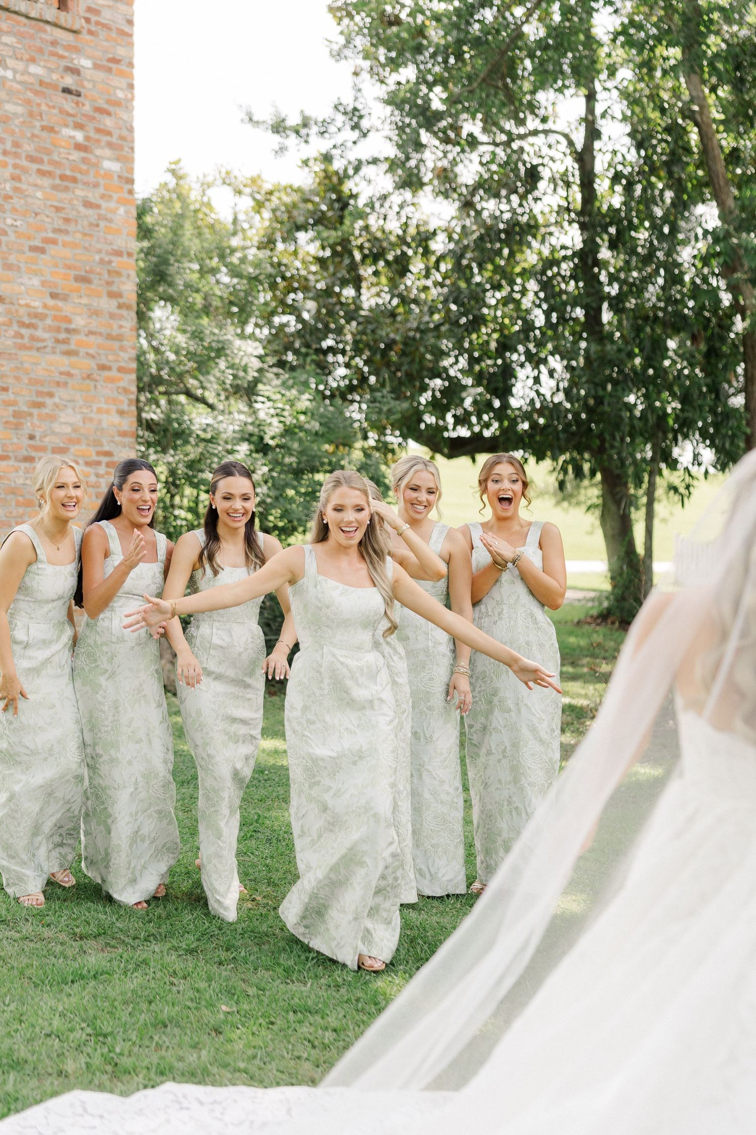 Six bridesmaids in sage green gowns react with excitement seeing the bride outdoors near brick building.