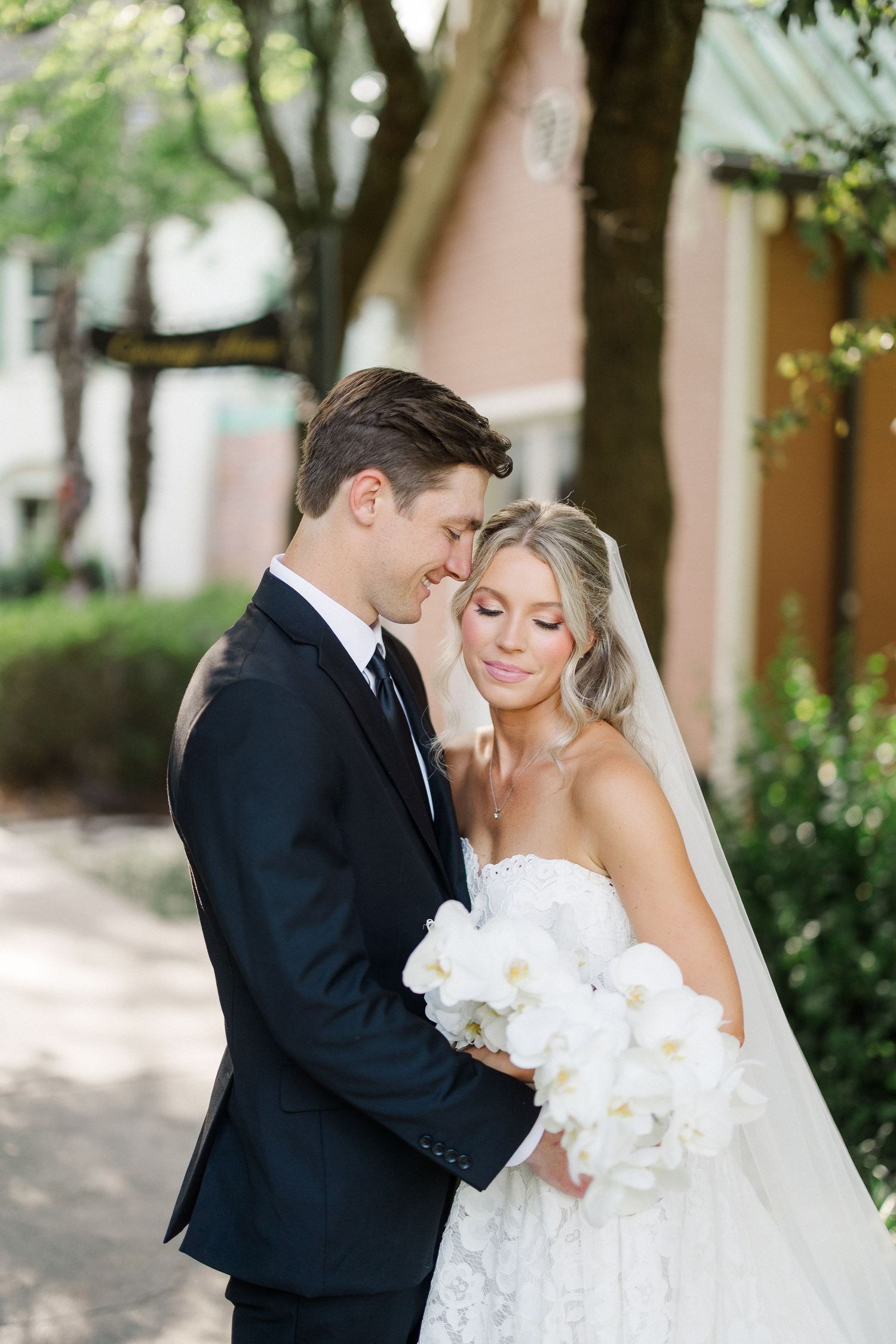 Bride and groom share a romantic moment outdoors, bride holding white orchid bouquet in lace gown with veil.