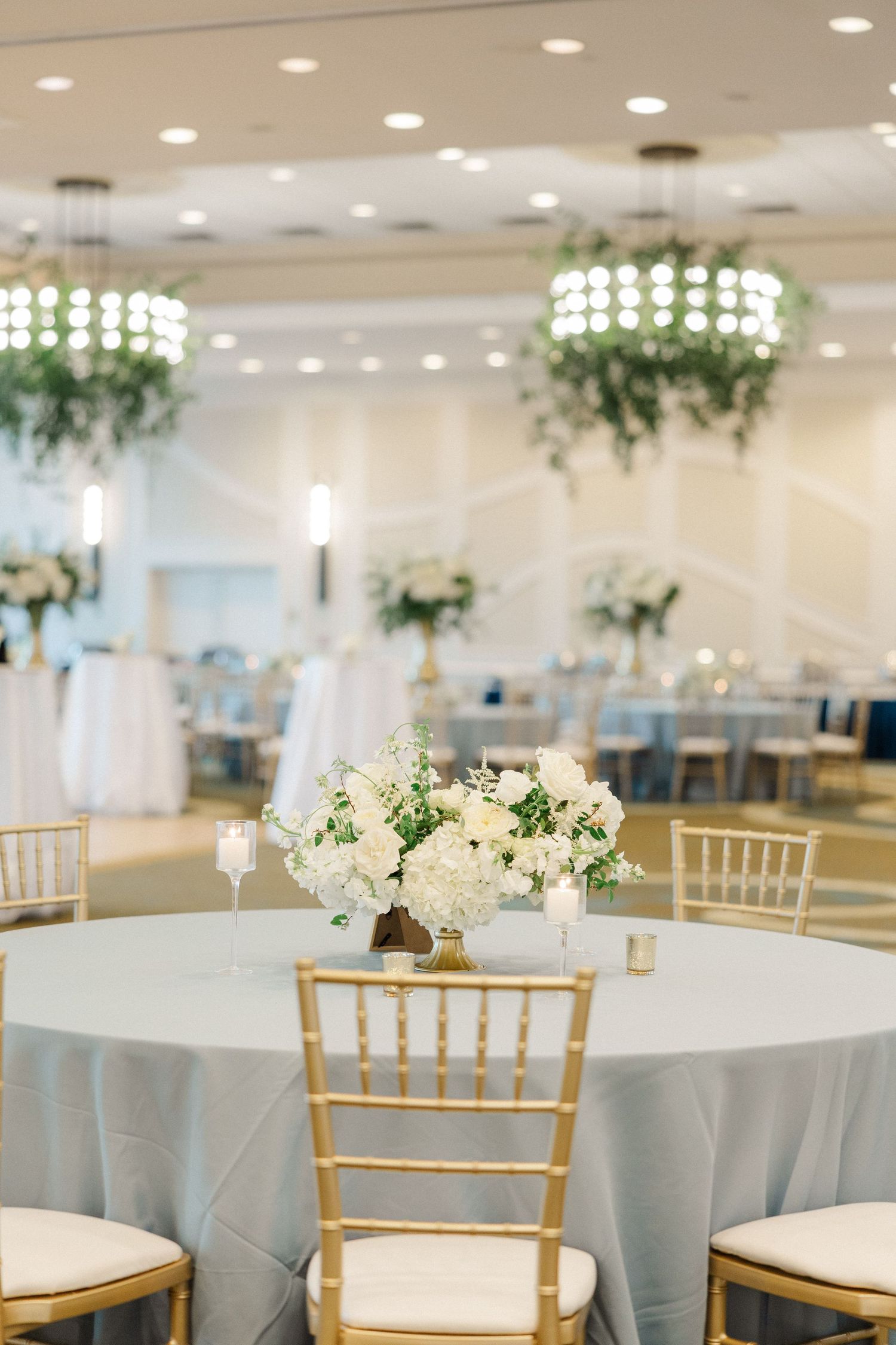 Elegant wedding reception with gold chiavari chairs, white floral centerpieces, and hanging greenery chandeliers in a bright ballroom.