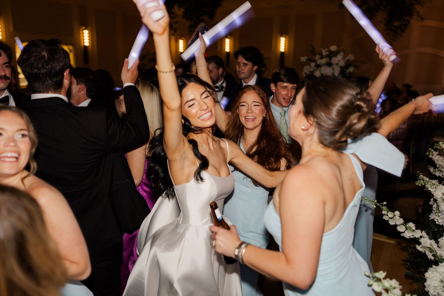 Joyful bride in white satin gown dancing with bridesmaids in light blue dresses at lively wedding reception.