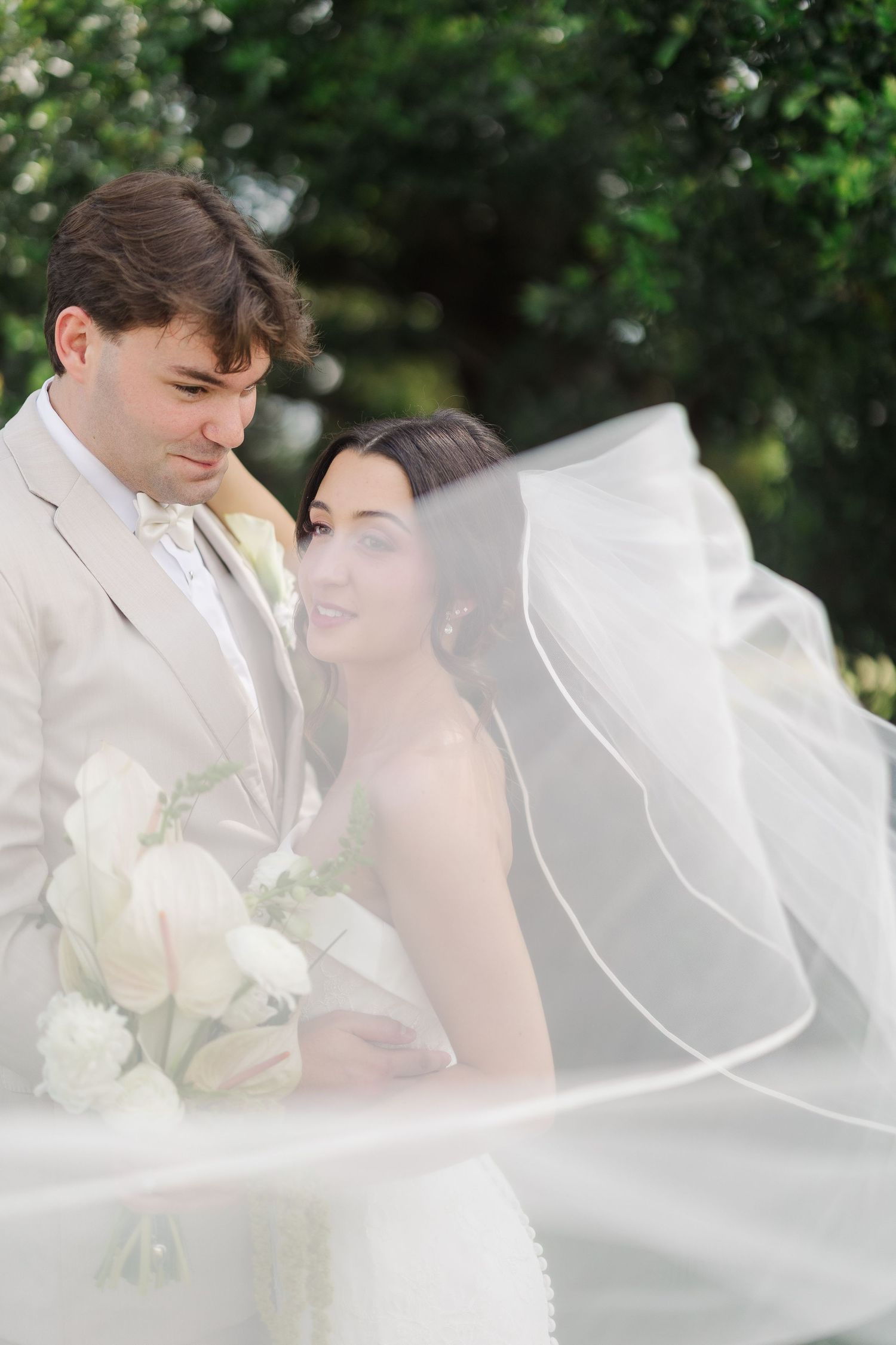 Romantic portrait of bride and groom wrapped in flowing veil among lush green garden backdrop.