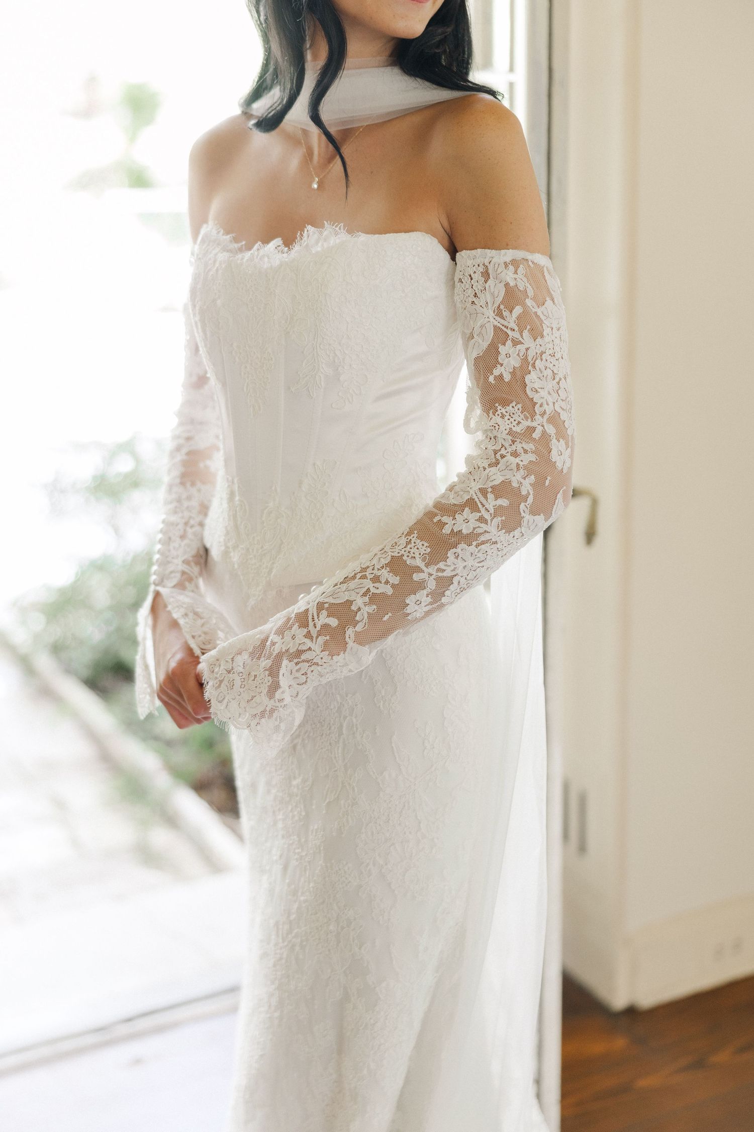Bride wearing an elegant off-shoulder white lace wedding gown with sheer long sleeves and fitted silhouette.