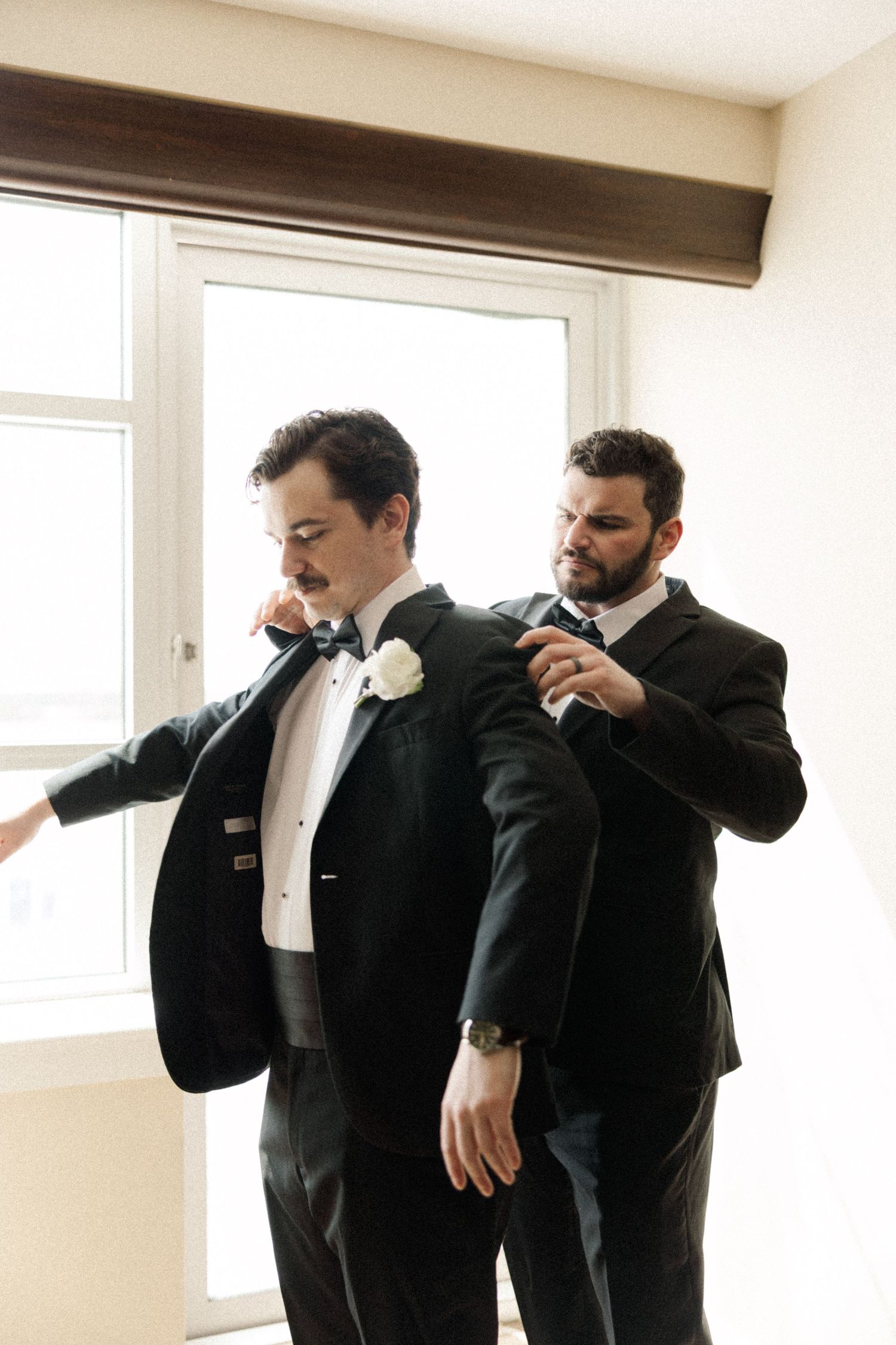 Groom being helped into his black tuxedo jacket by groomsman before wedding ceremony.