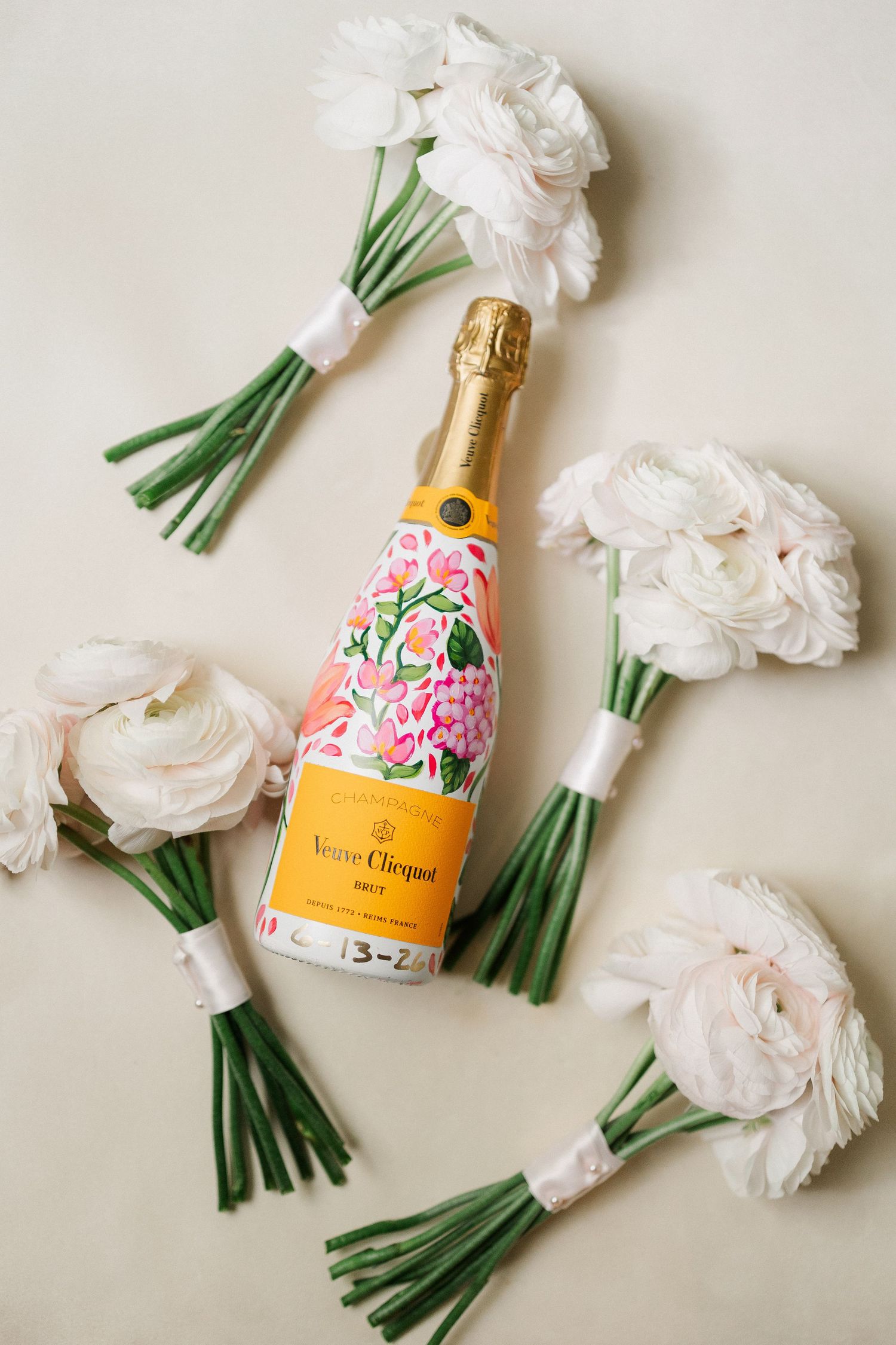 Veuve Cliquot Brut champagne bottle with floral label surrounded by white ranunculus bouquets on cream background.
