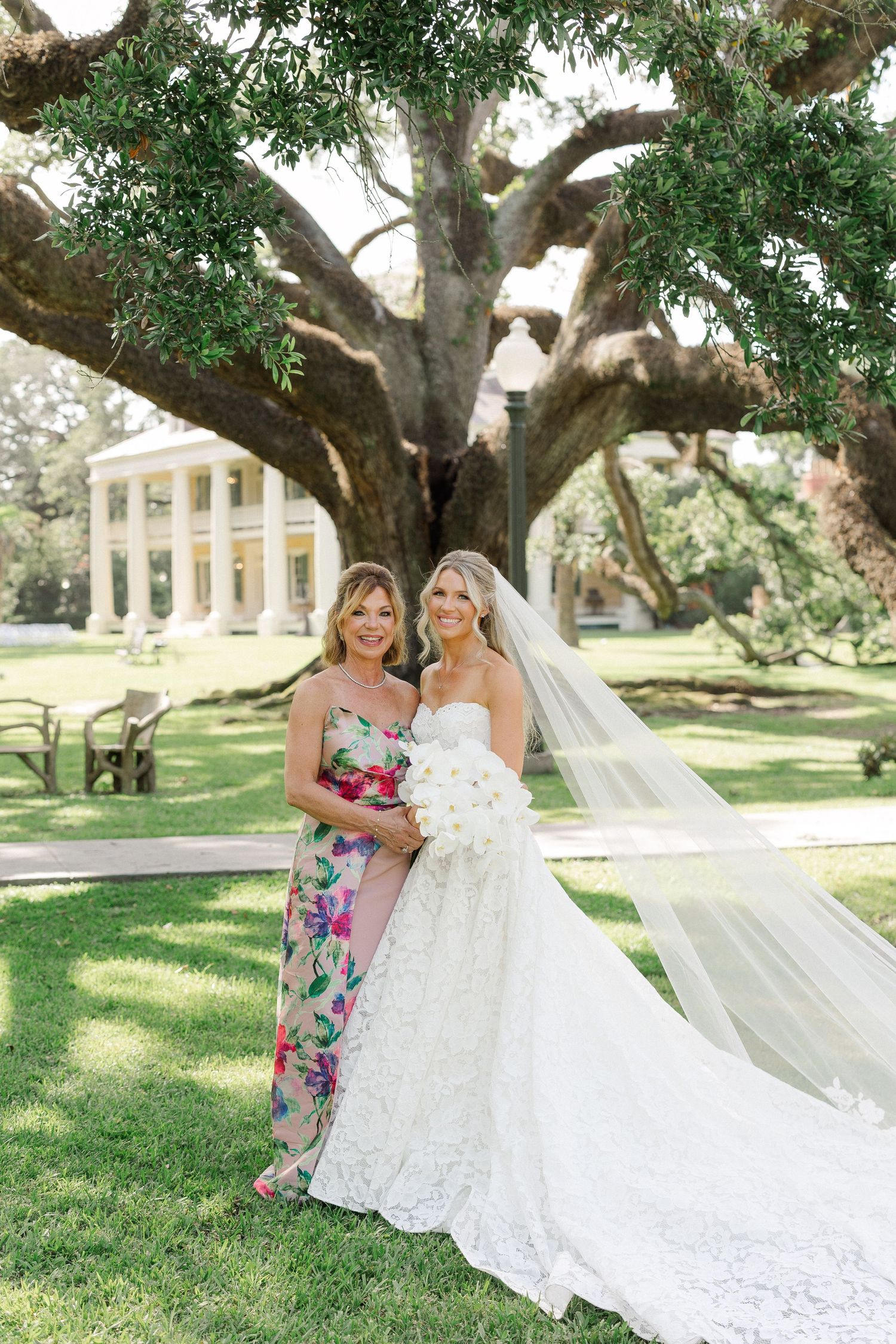 Bride in strapless lace gown with veil poses with mother in floral dress under grand oak tree.