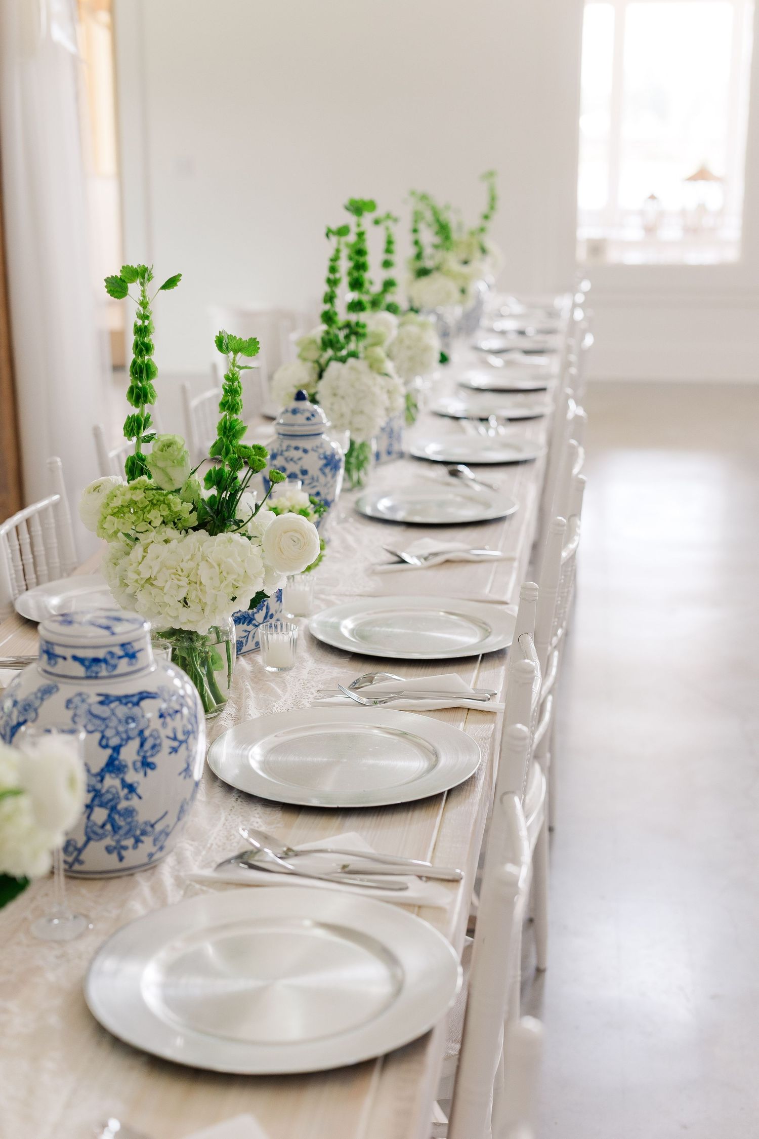 Elegant long dining table with white plates, silver chargers, blue and white vases, and green floral arrangements.