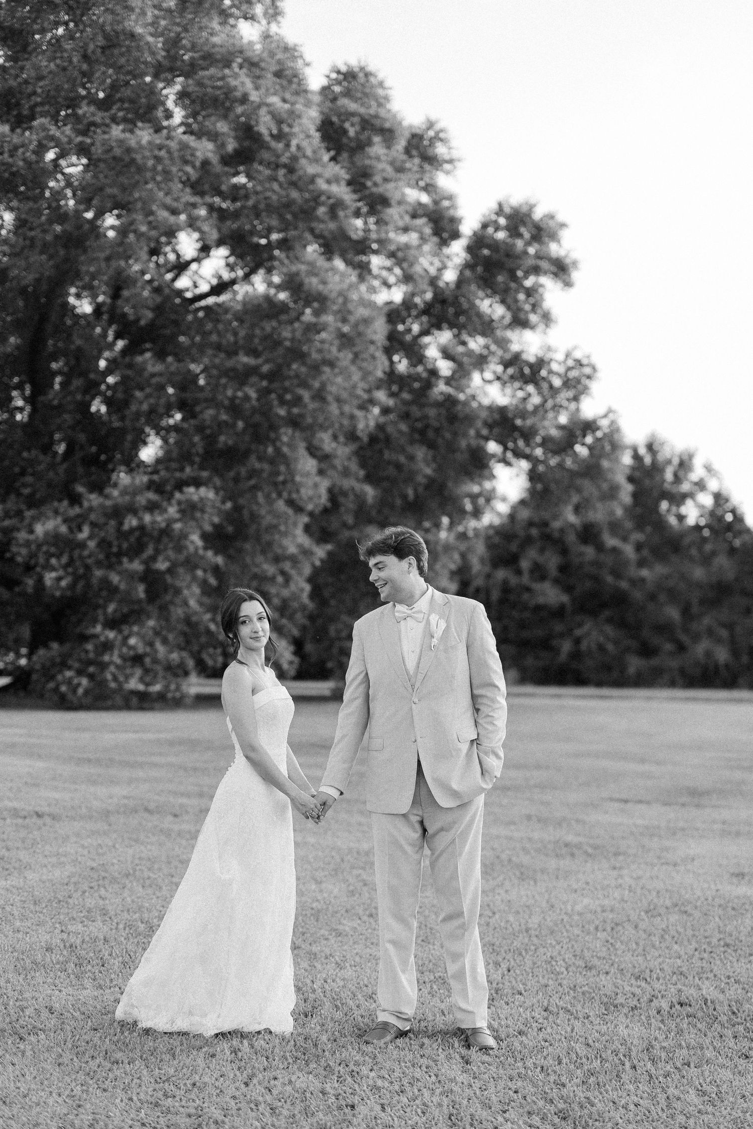 Black and white portrait of bride and groom holding hands in open green field surrounded by trees.