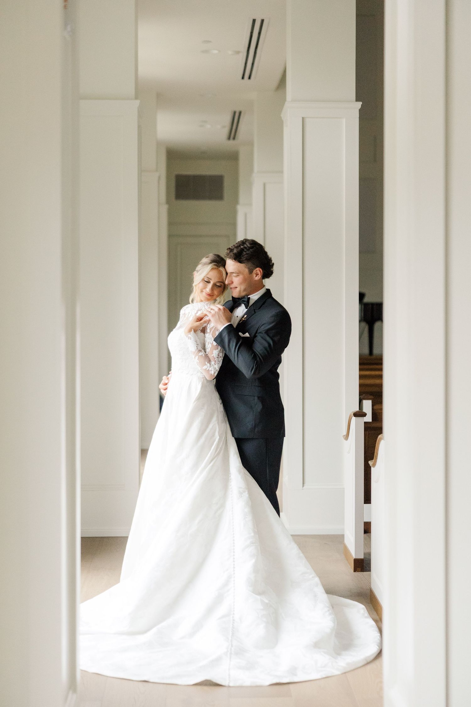 A bride and groom share a tender embrace in a bright white hallway, the bride wearing a long lace wedding gown with a train.