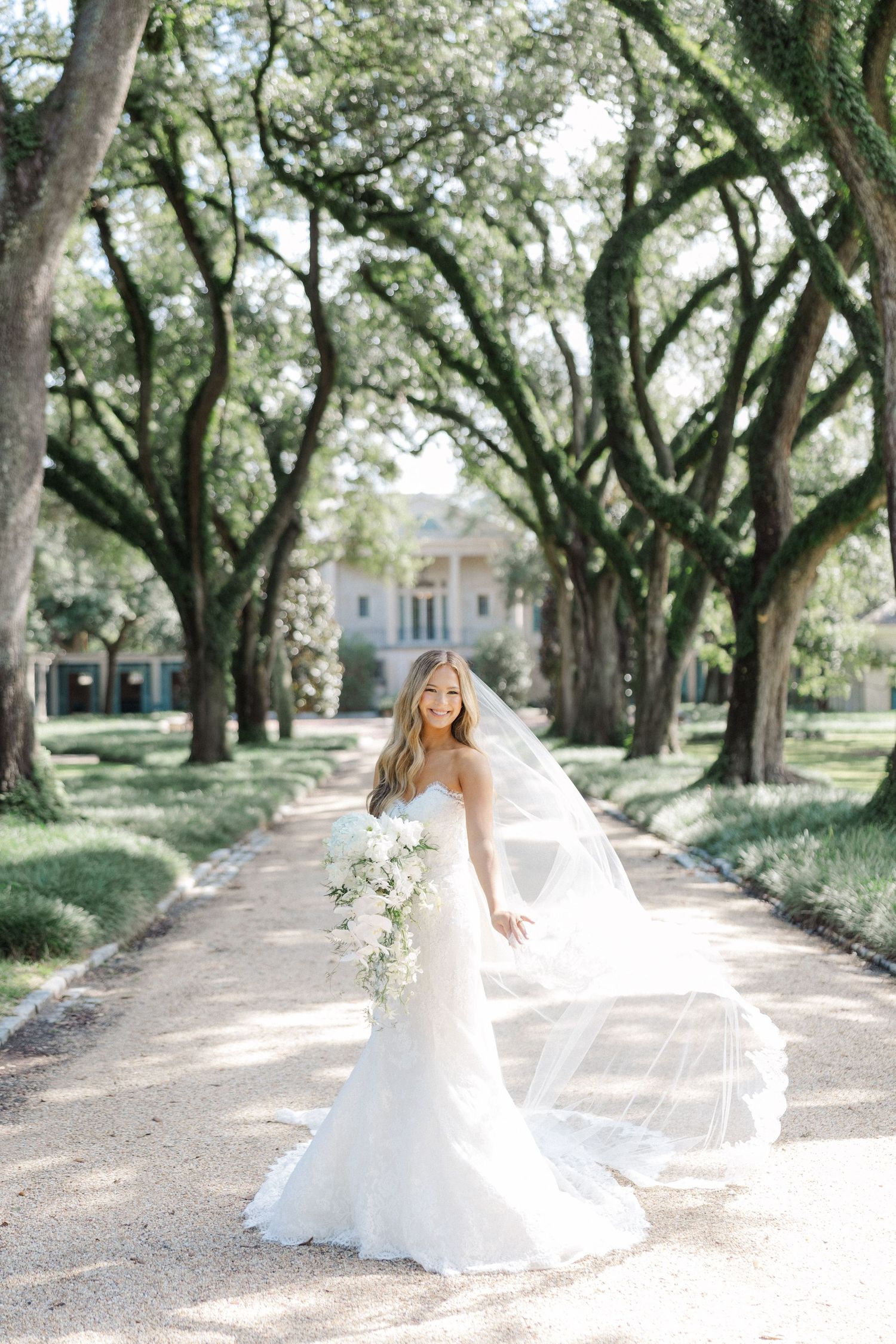 Elegant bridal portrait under a canopy of oak trees with a white mansion visible in the background.
