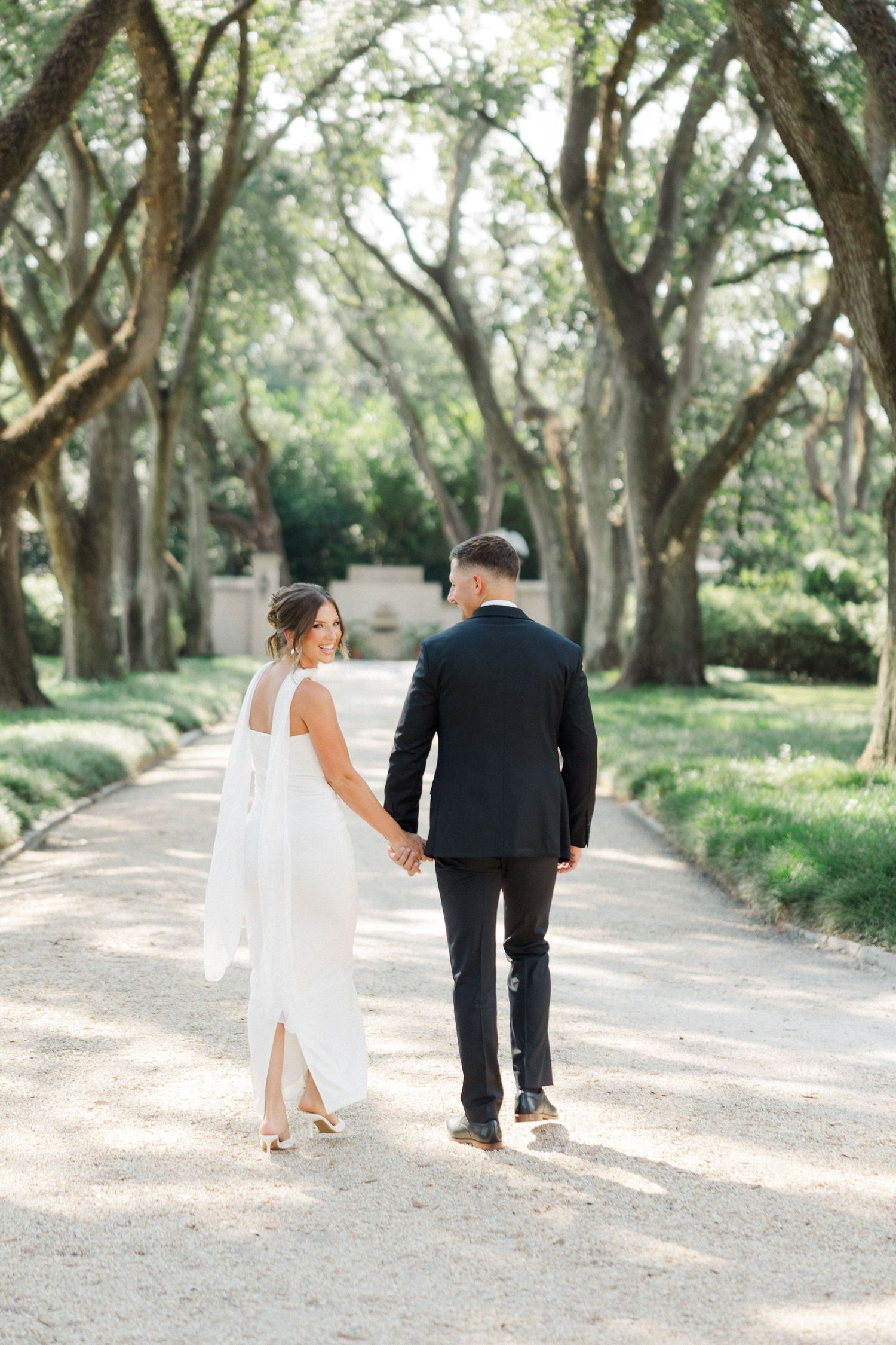 Newlywed couple walking down a tree-lined path in a romantic garden setting.