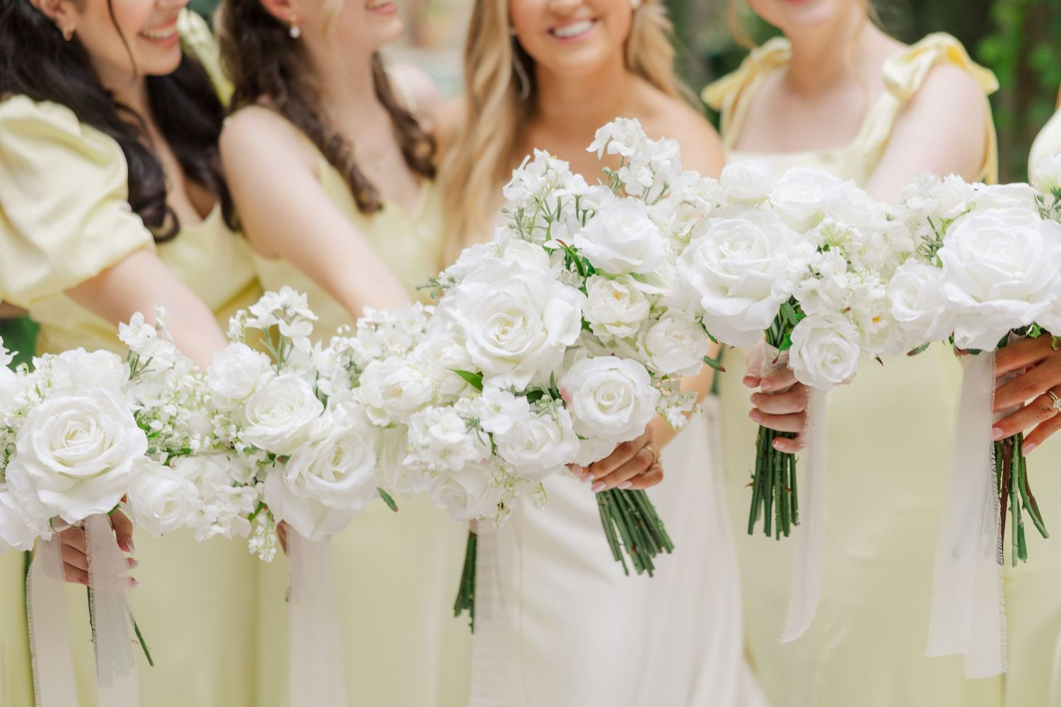 Row of white floral wedding bouquets held by bridesmaids in formal attire against outdoor garden backdrop.