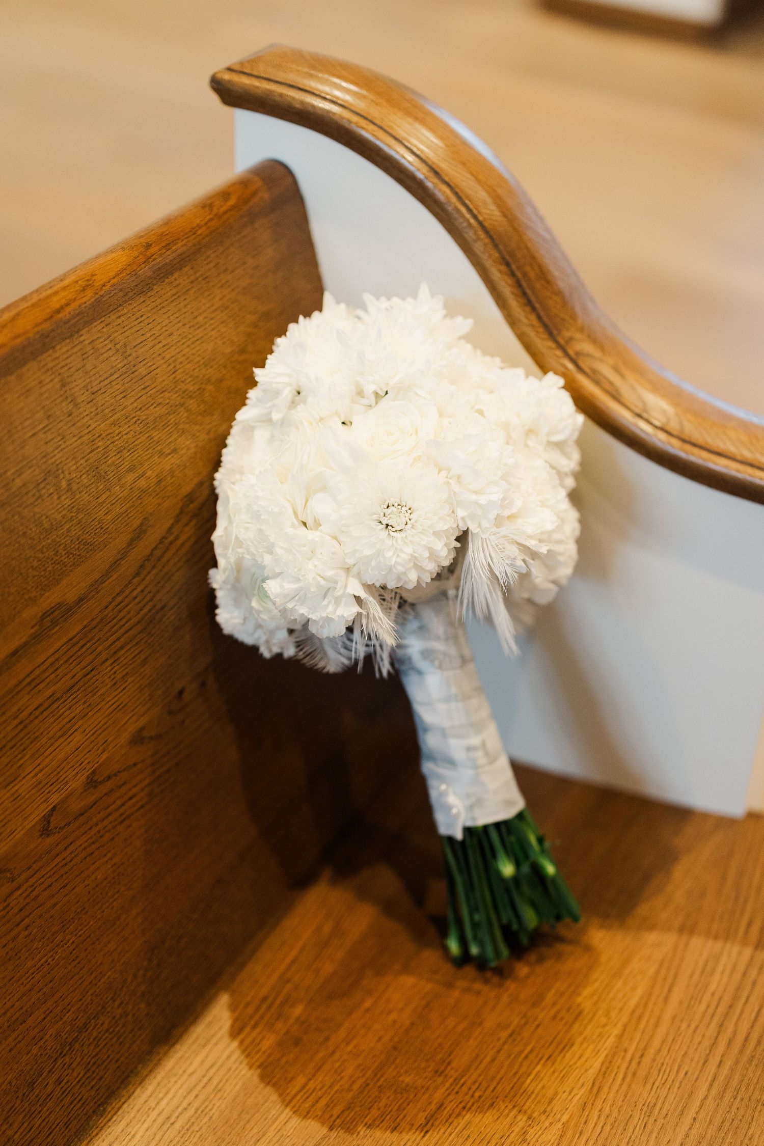 A white bridal bouquet with delicate carnations rests on a polished wooden surface, wrapped in a satin ribbon.