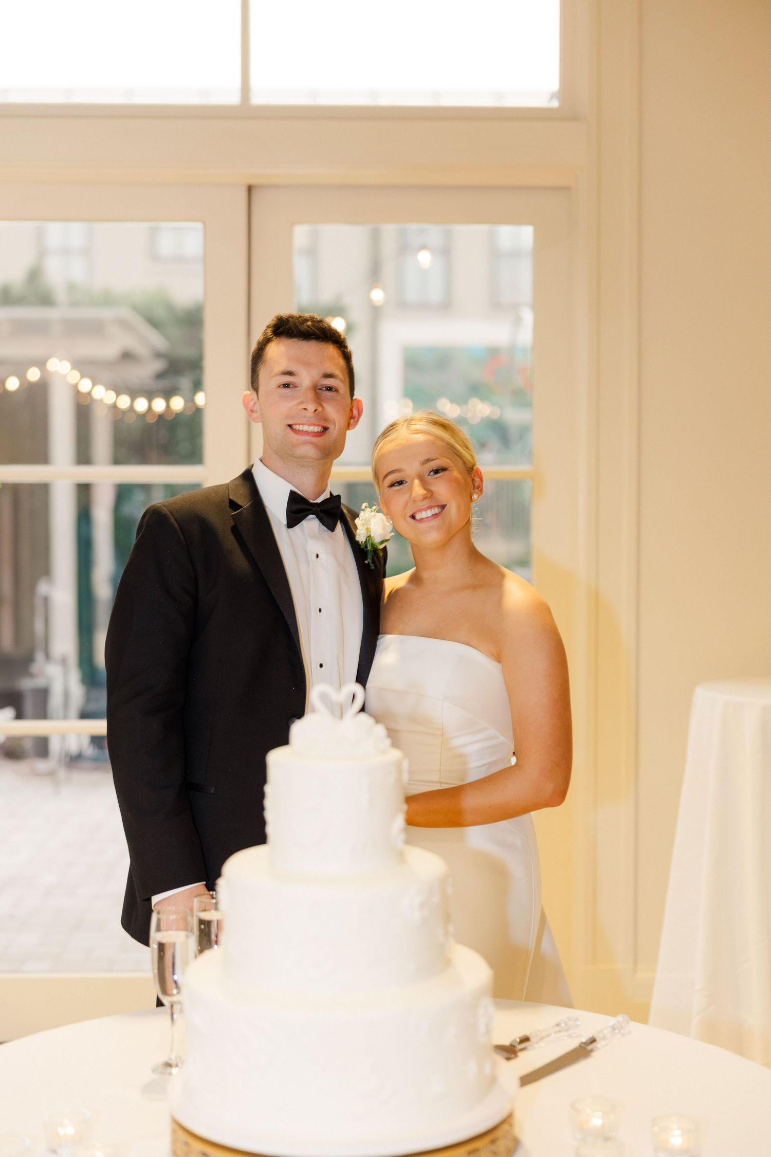 A wedding couple poses together next to their elegant white tiered wedding cake.