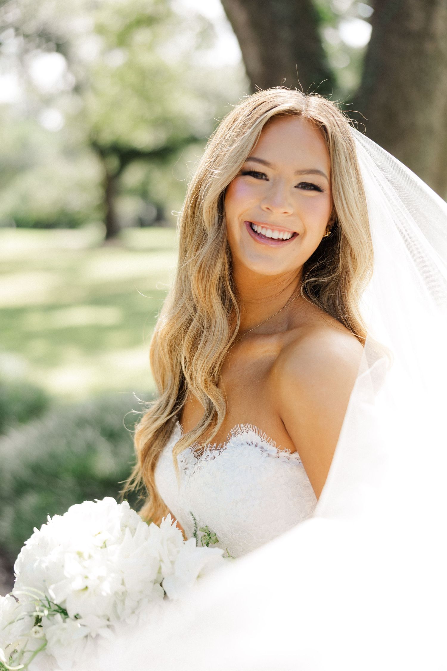 A bride in a white wedding dress holds a bouquet of white flowers while smiling in an outdoor garden setting.