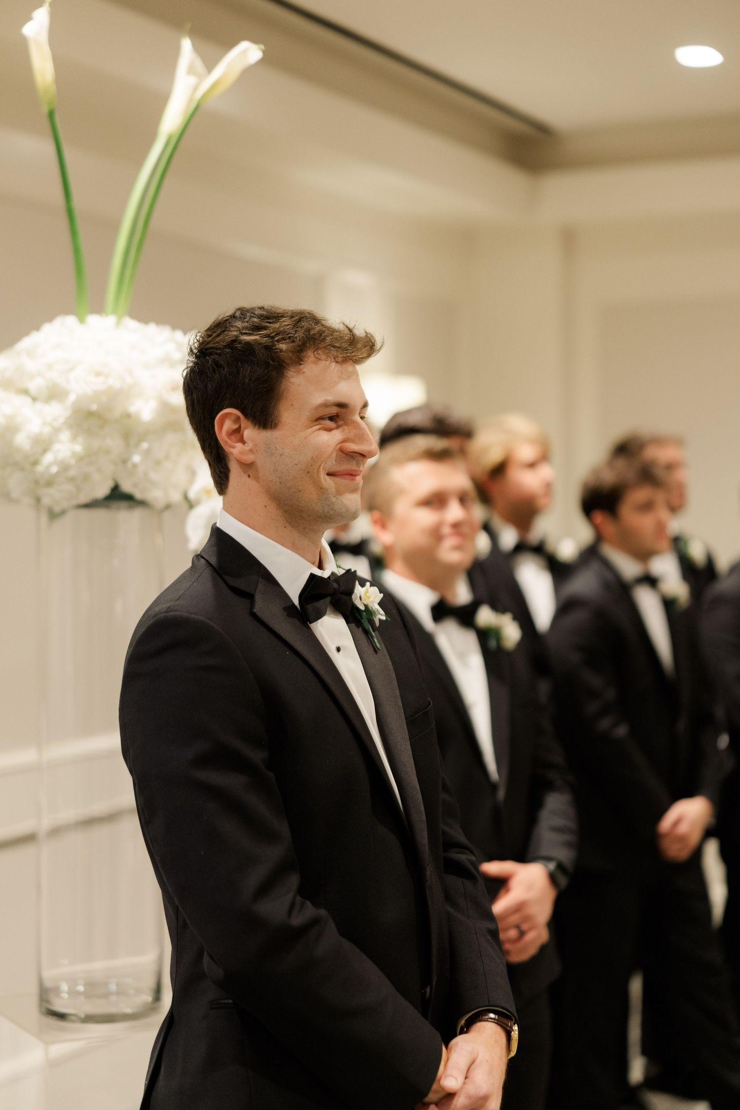 Groomsmen in black tuxedos lined up at an elegant indoor wedding ceremony.