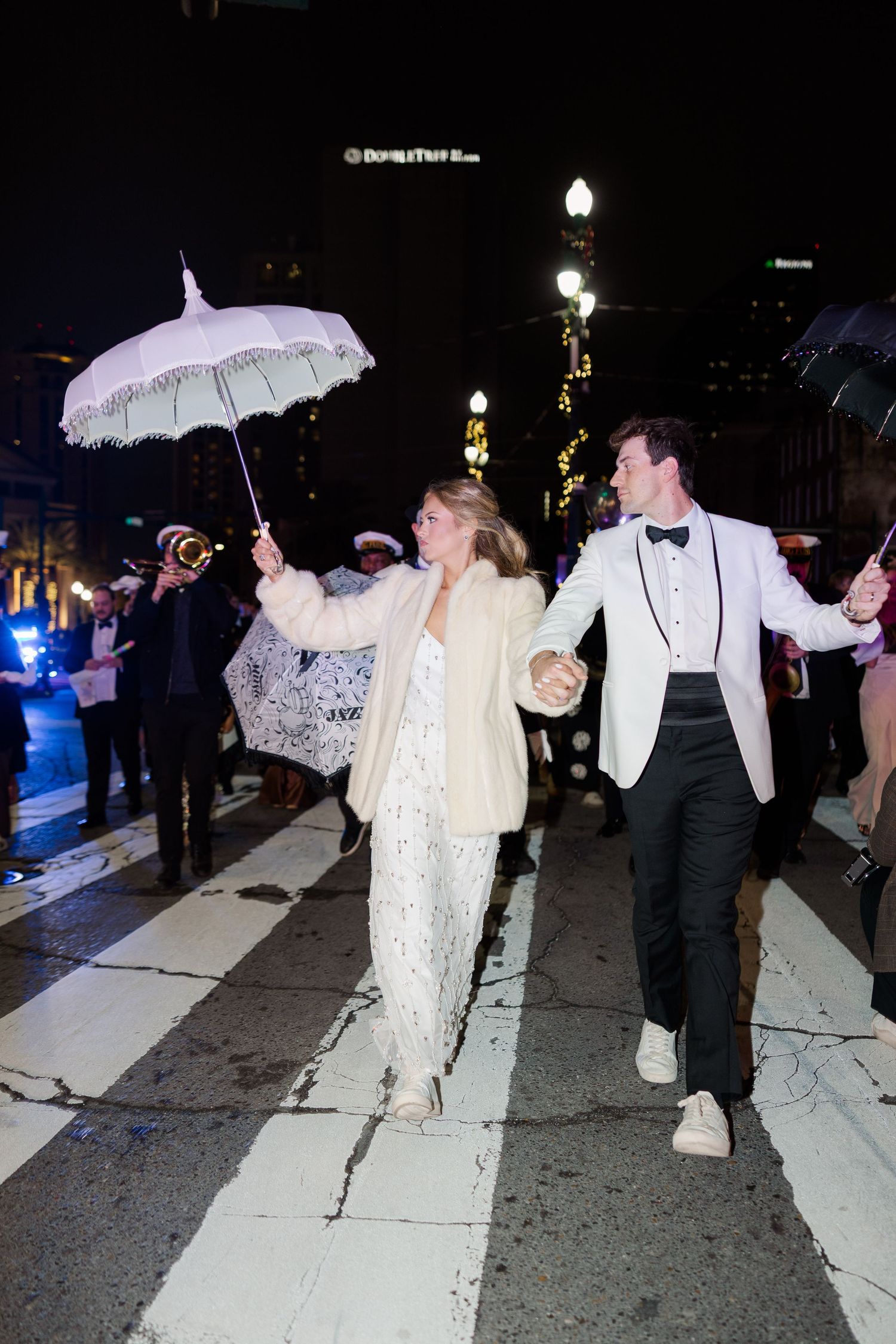 Couple walks down city street at night holding a decorative white parasol during wedding celebration.