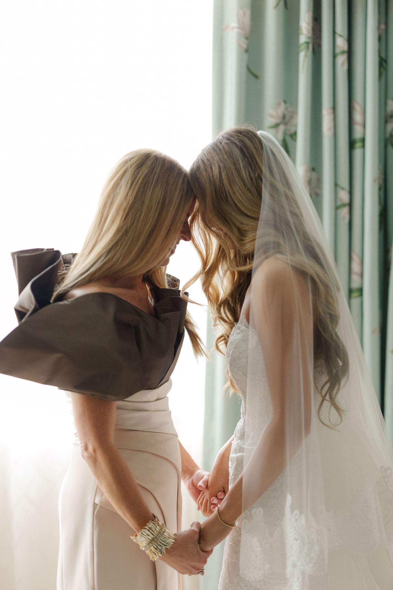 Two people share an intimate moment in formal wedding attire near sheer curtains.