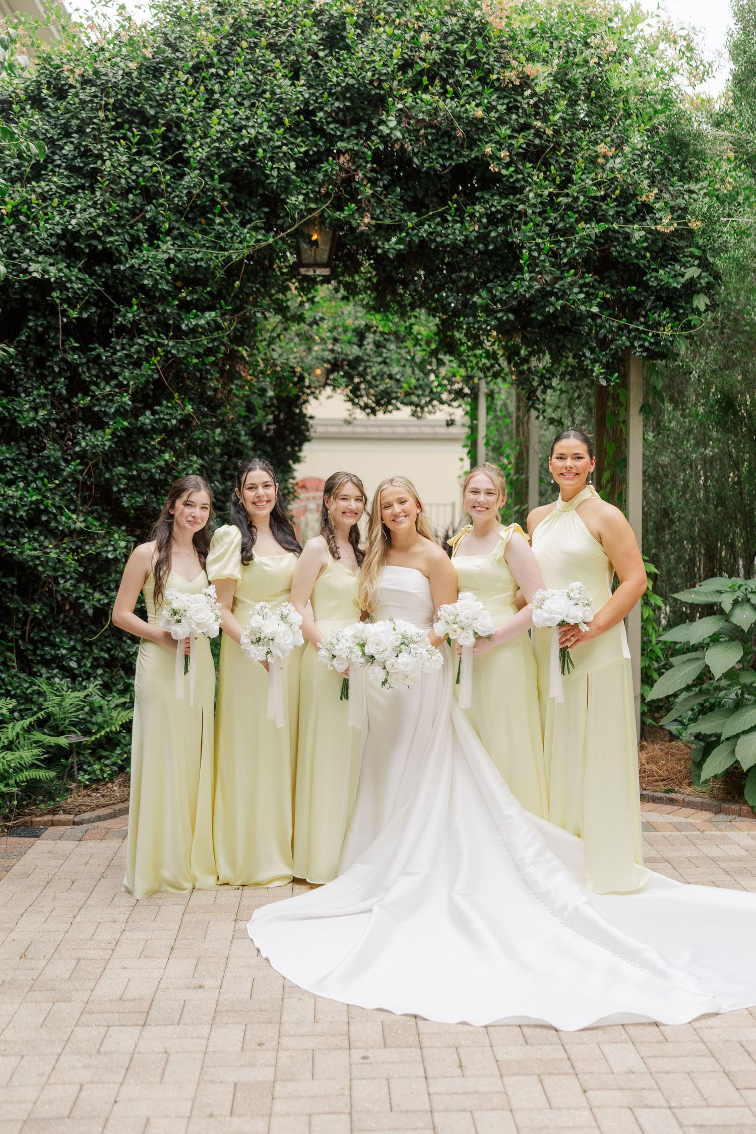 Bridesmaids in soft yellow dresses pose with the bride in a white gown against a lush garden backdrop.