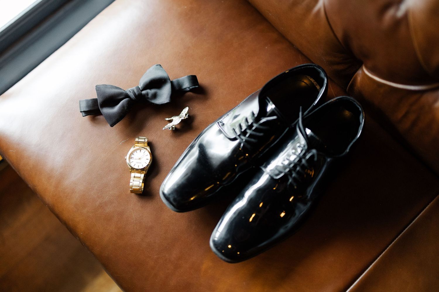 A pair of shiny black leather dress shoes sits on a brown leather surface in warm lighting.