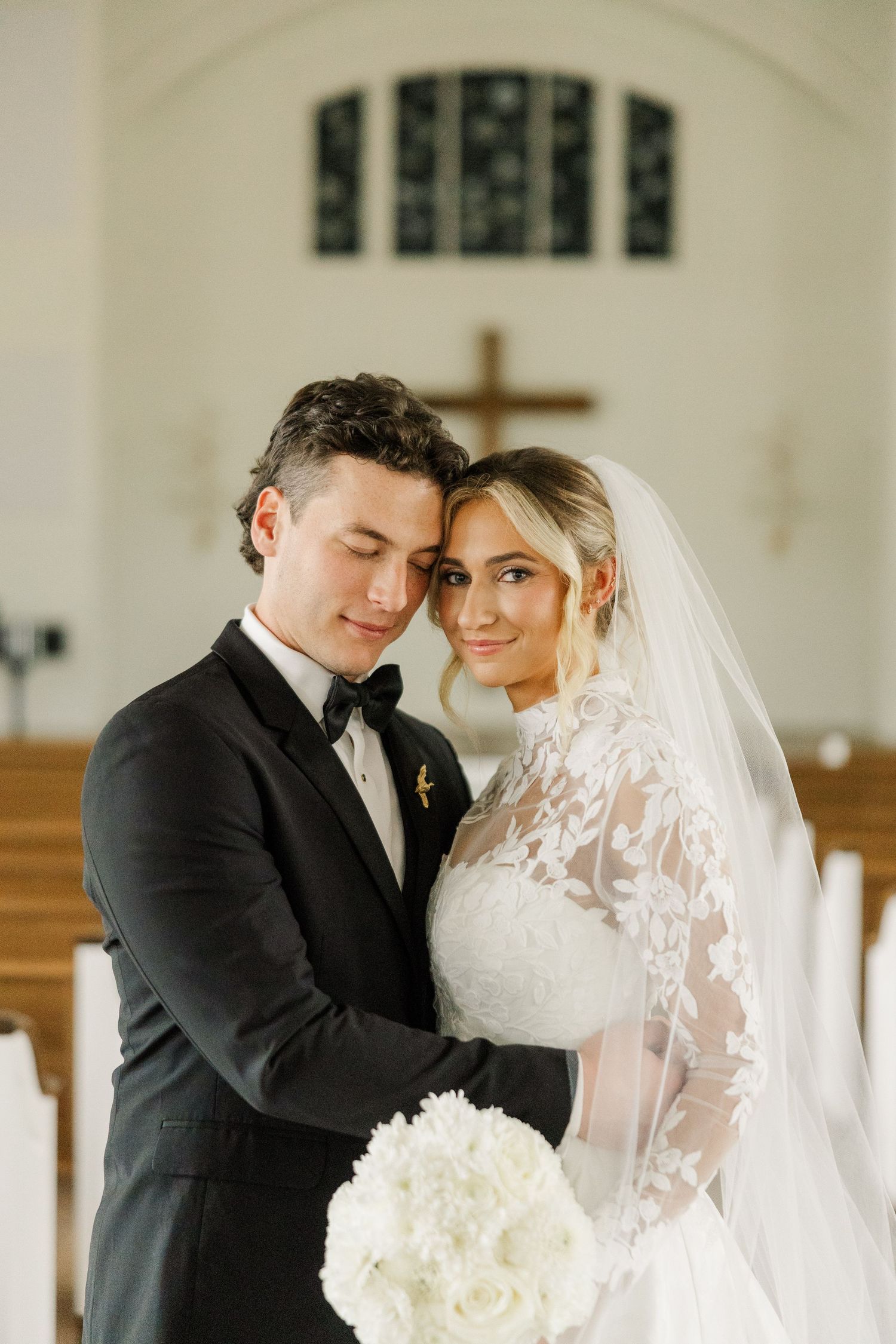 A tender wedding moment captured in a church with white floral bouquet and lace veil details.