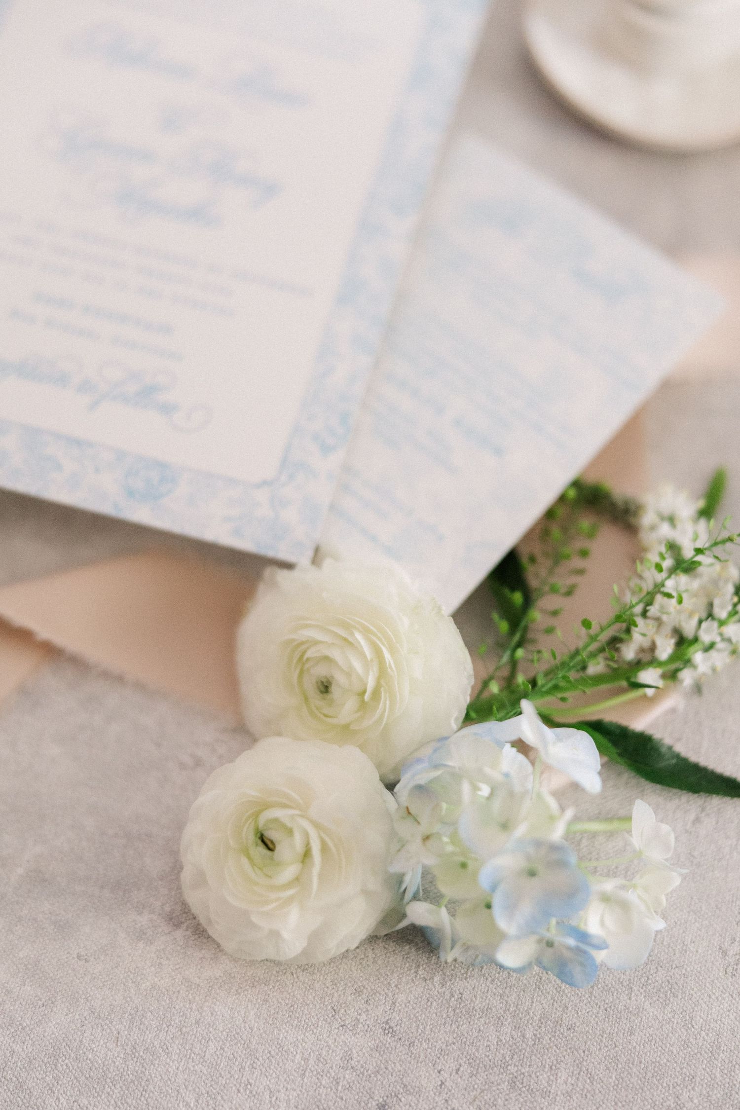 Delicate white ranunculus flowers and blue hydrangea blooms arranged with wedding stationery on a light gray surface.