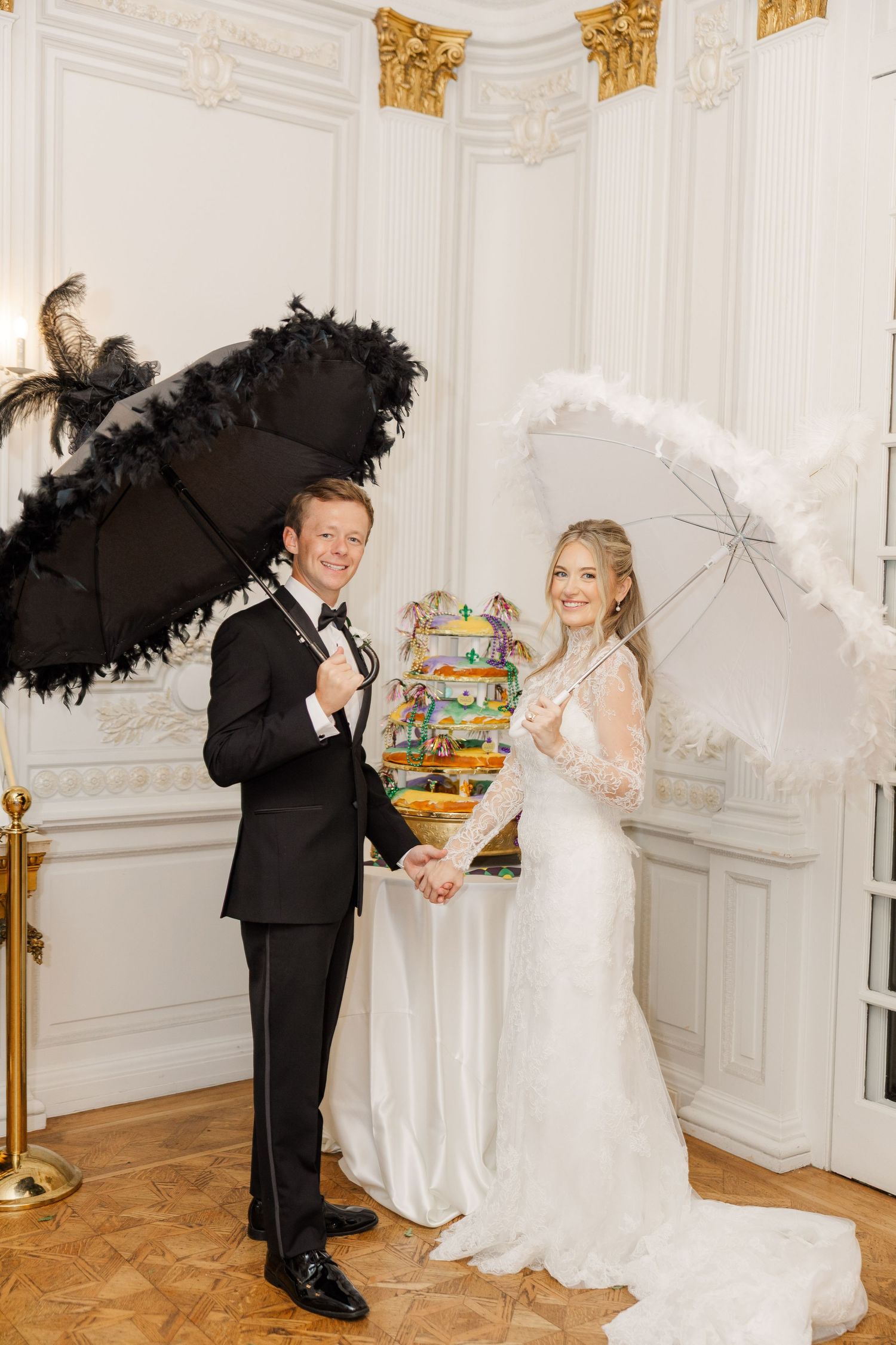 Wedding couple poses playfully with parasols in an elegant white and gold decorated room.