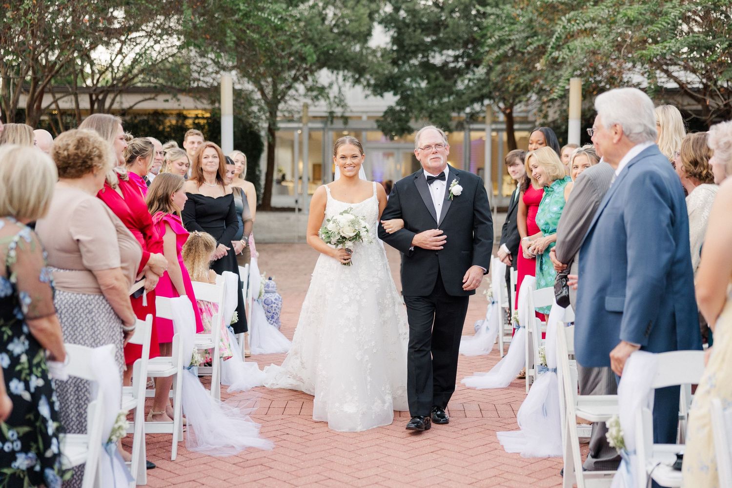 Wedding ceremony in progress with guests seated in white chairs on brick pathway.