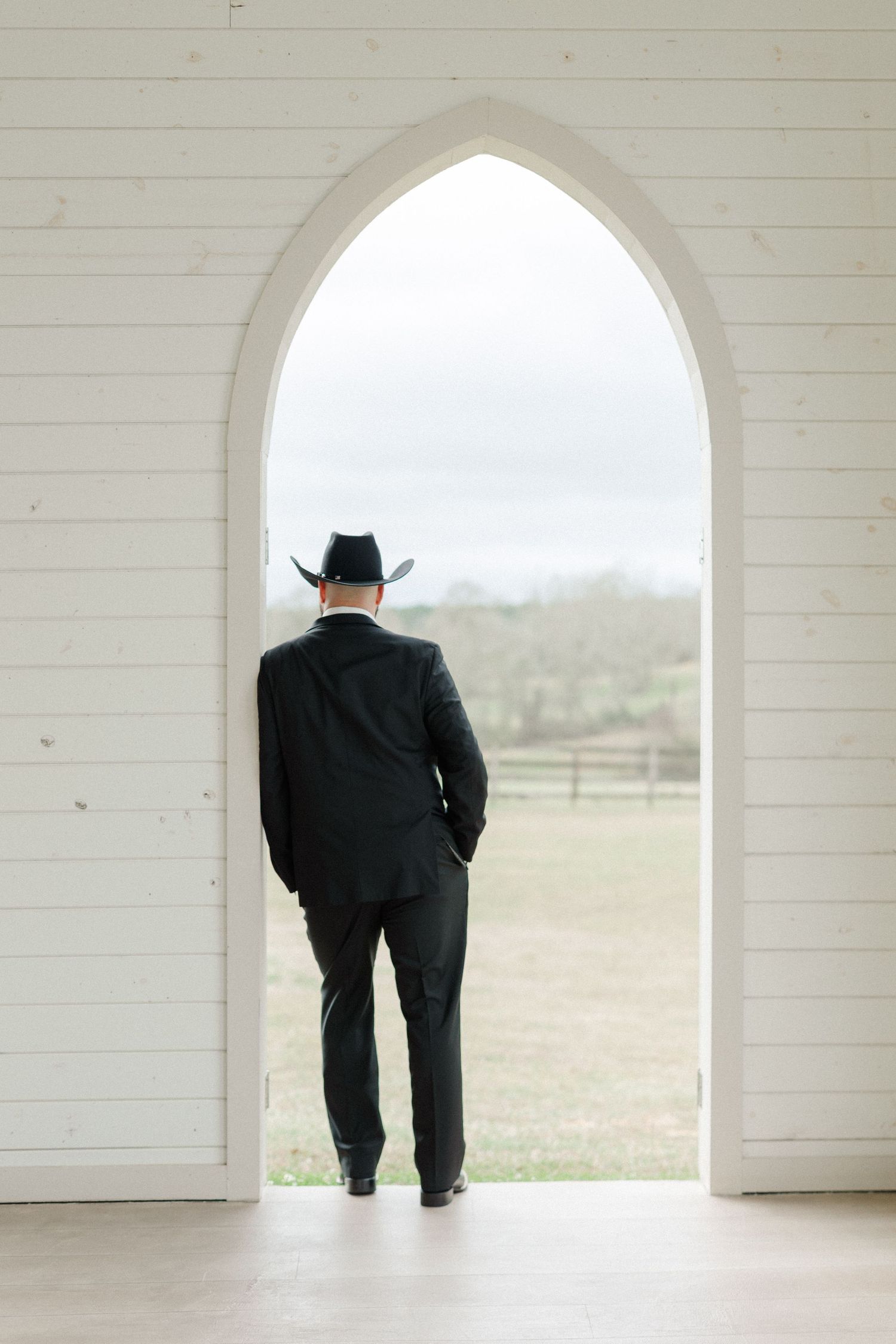 A person in a black suit and cowboy hat stands silhouetted in a white gothic archway looking out at rural landscape.