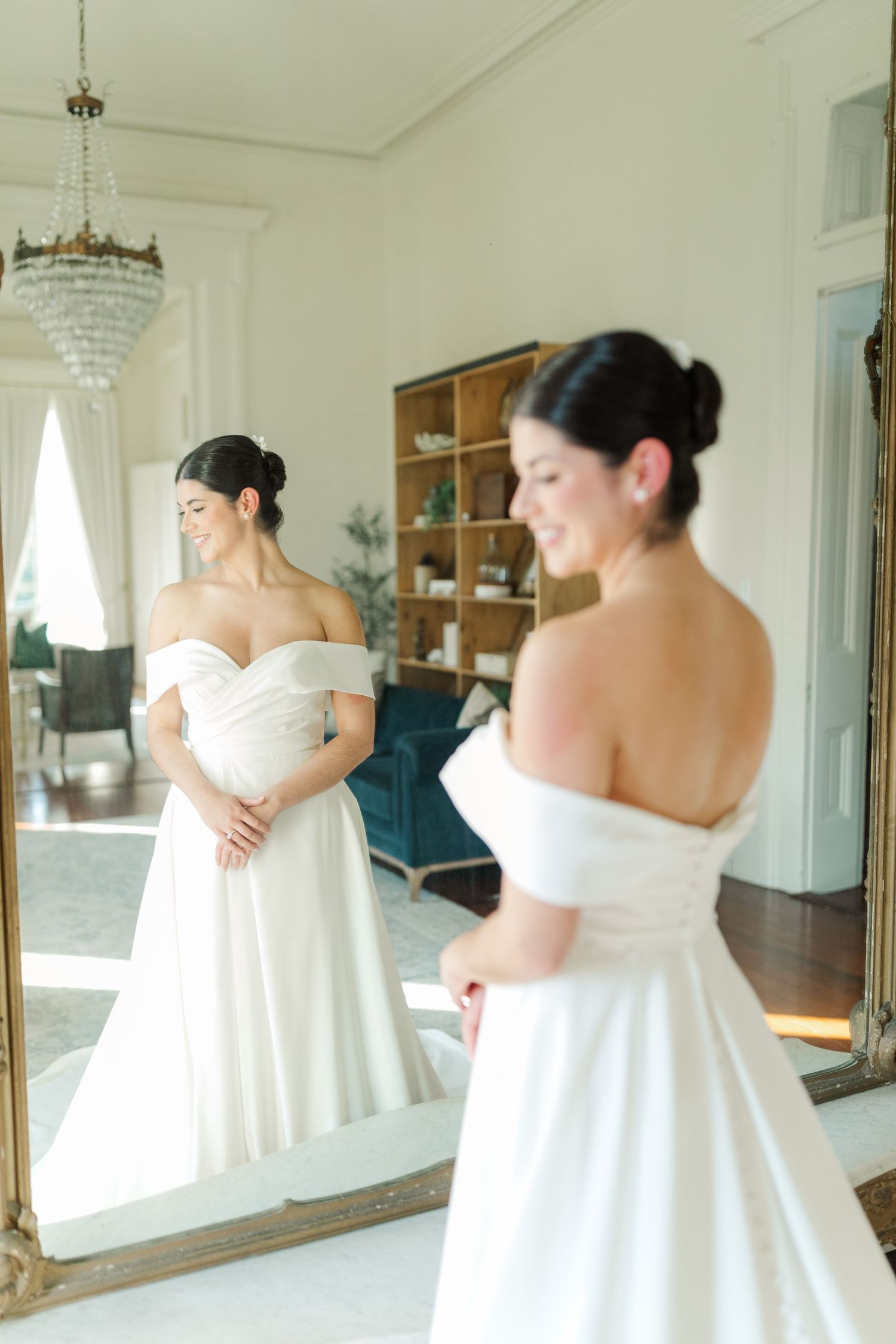 An elegant off-shoulder white wedding dress is shown being fitted in a bright room with a crystal chandelier and mirror.