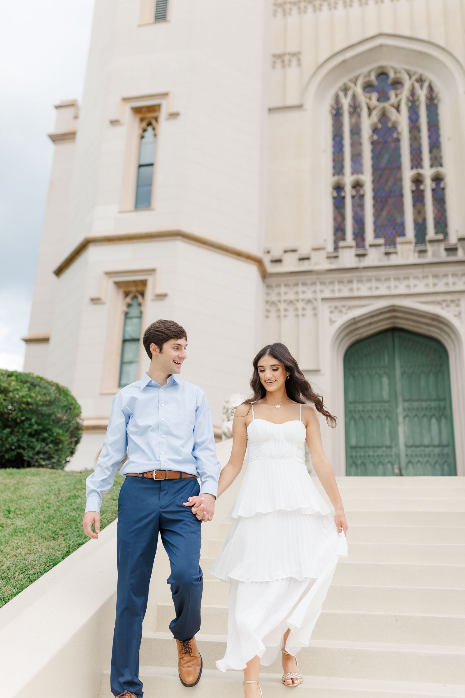 A couple in wedding attire walk down stone steps in front of a gothic-style cathedral with tall stained glass windows.