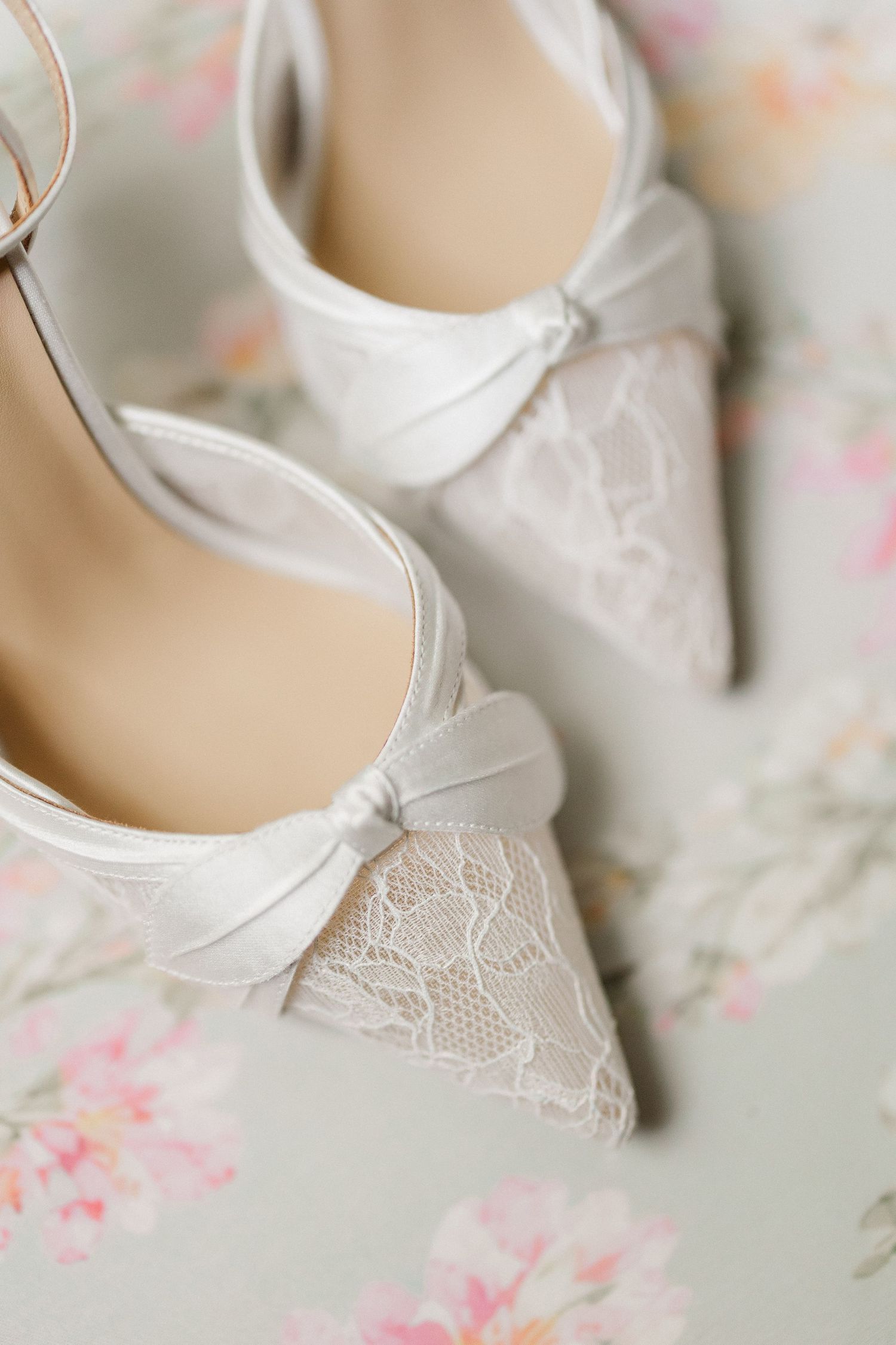 Elegant white bridal heels with lace toe detail and satin bow on floral background.