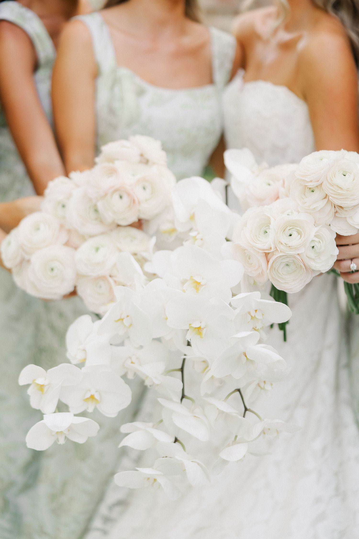 Bride and bridesmaids holding white orchid cascade and blush ranunculus bouquets in sage green dresses.