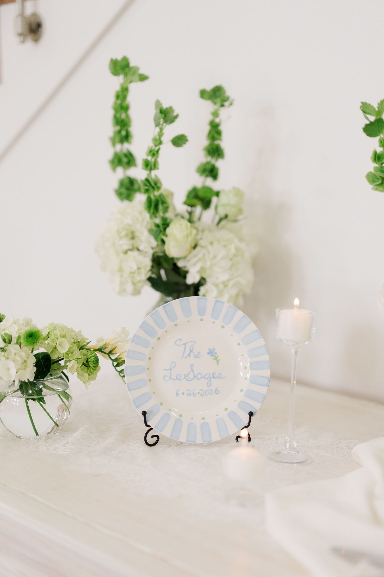 Decorative wedding plate on easel with blue stripe border, candle, and white green floral arrangements on white table.