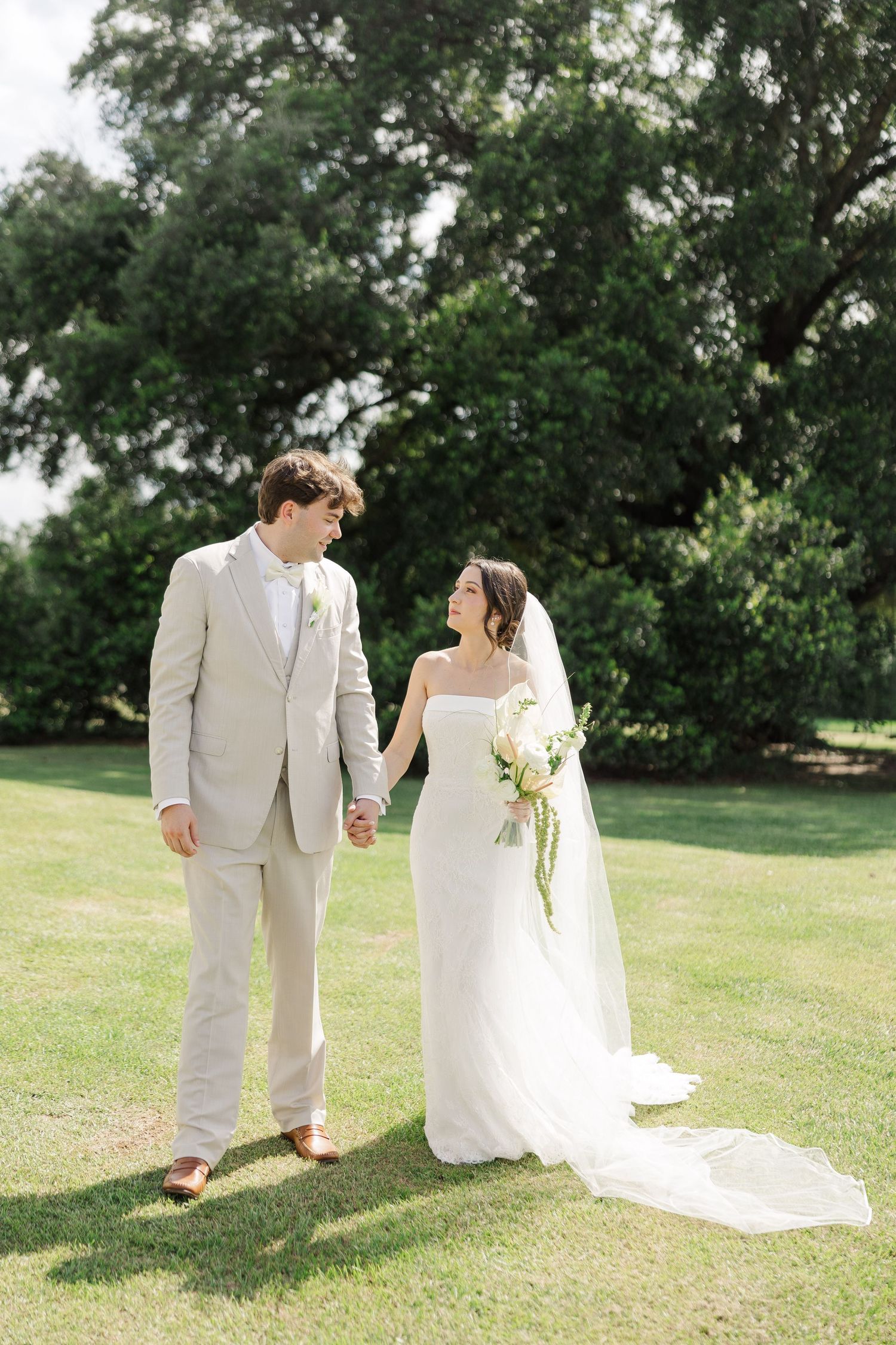 Bride and groom holding hands on sunlit green lawn, bride carrying white bouquet with long veil.