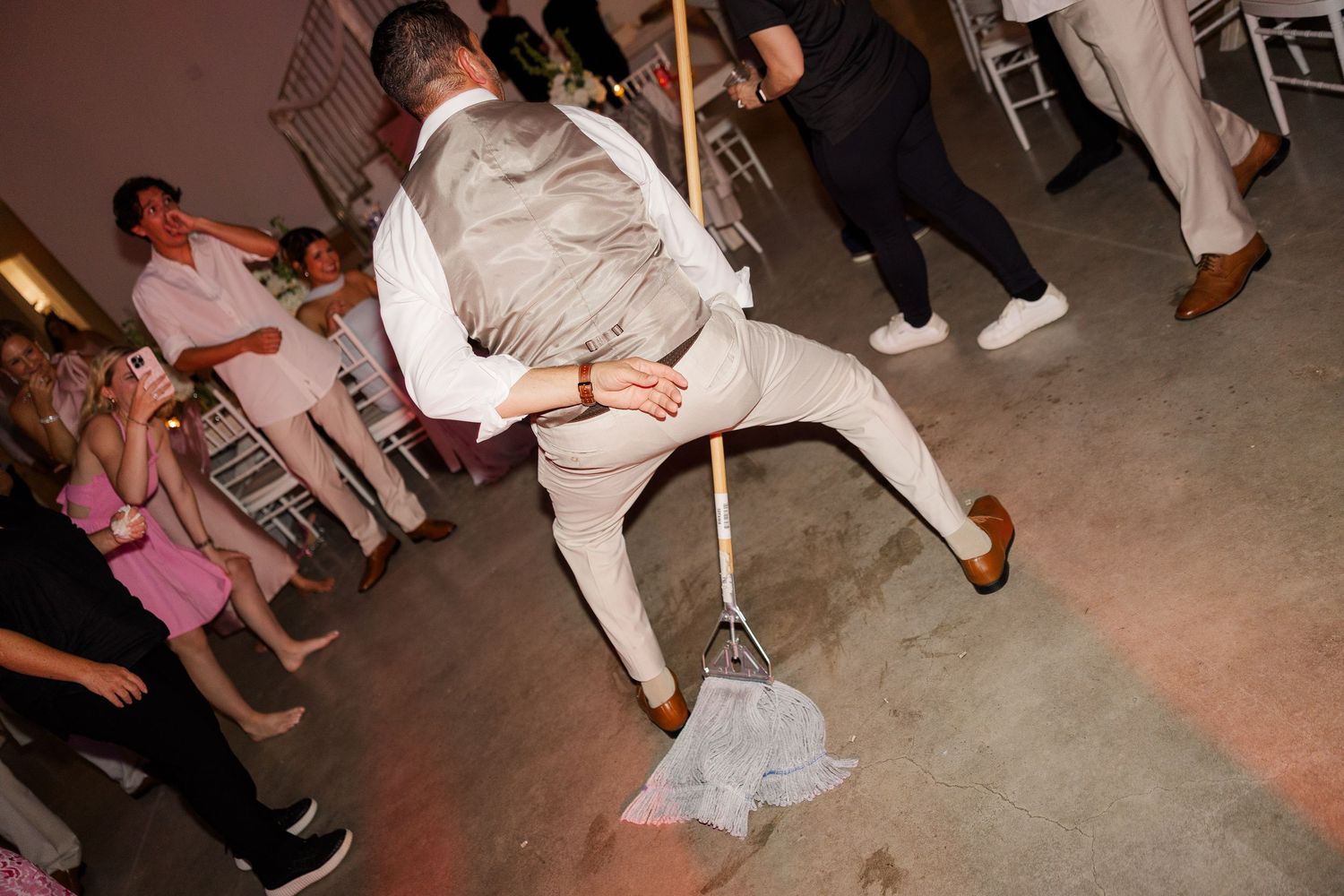 Groom in tan suit does limbo on wedding reception dance floor surrounded by cheering guests.