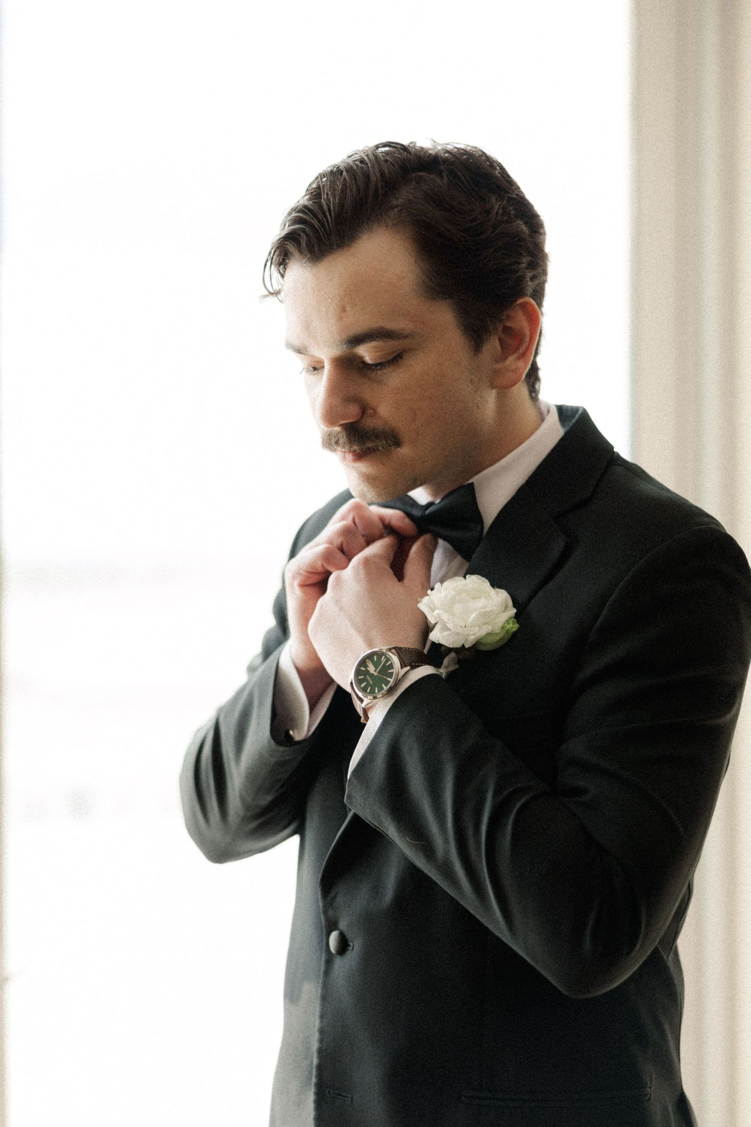 Dapper groom in black tuxedo adjusting bow tie with white rose boutonniere near bright window.