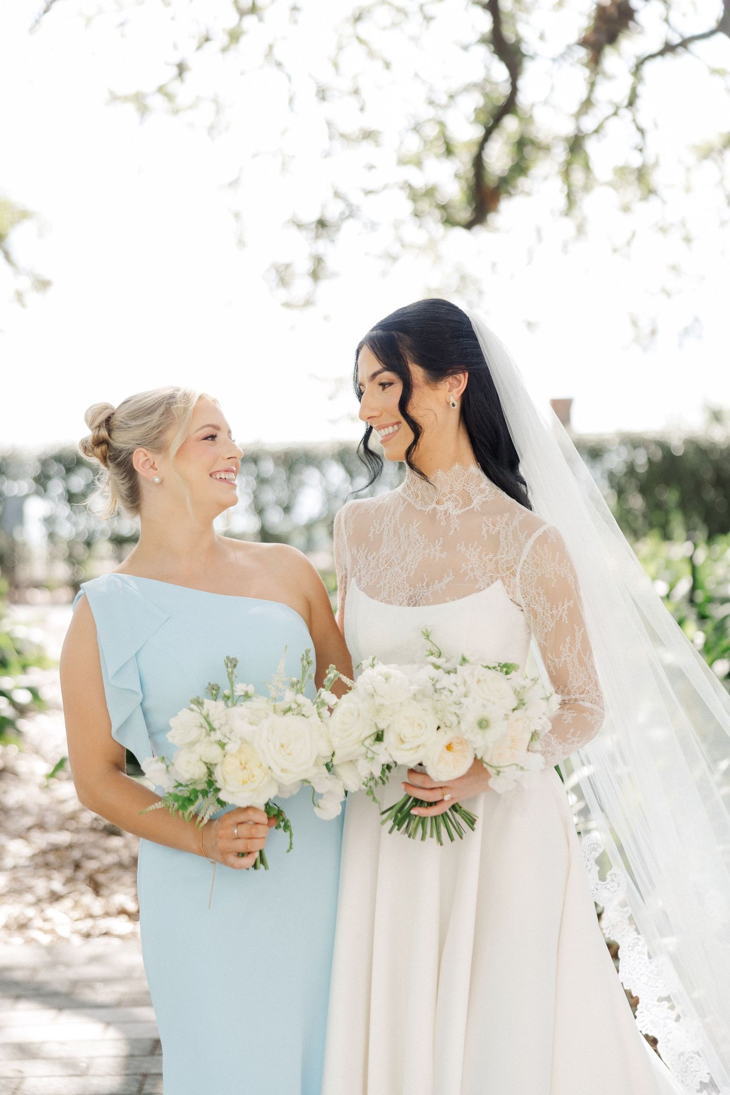 Bride in lace wedding gown and bridesmaid in light blue dress smile at each other holding white bouquets.