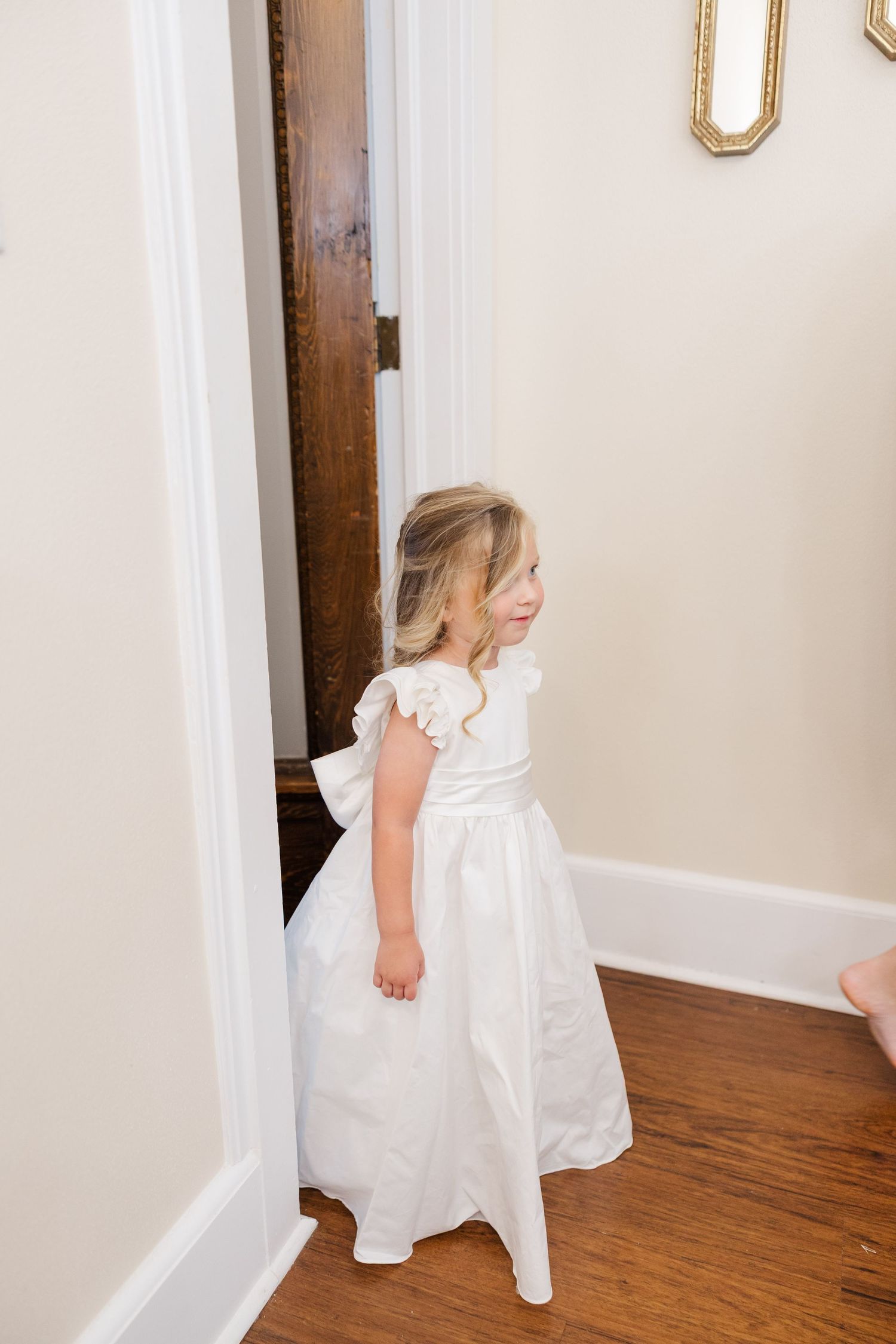 A young child in a white formal dress stands near a wooden door in a hallway with hardwood floors.