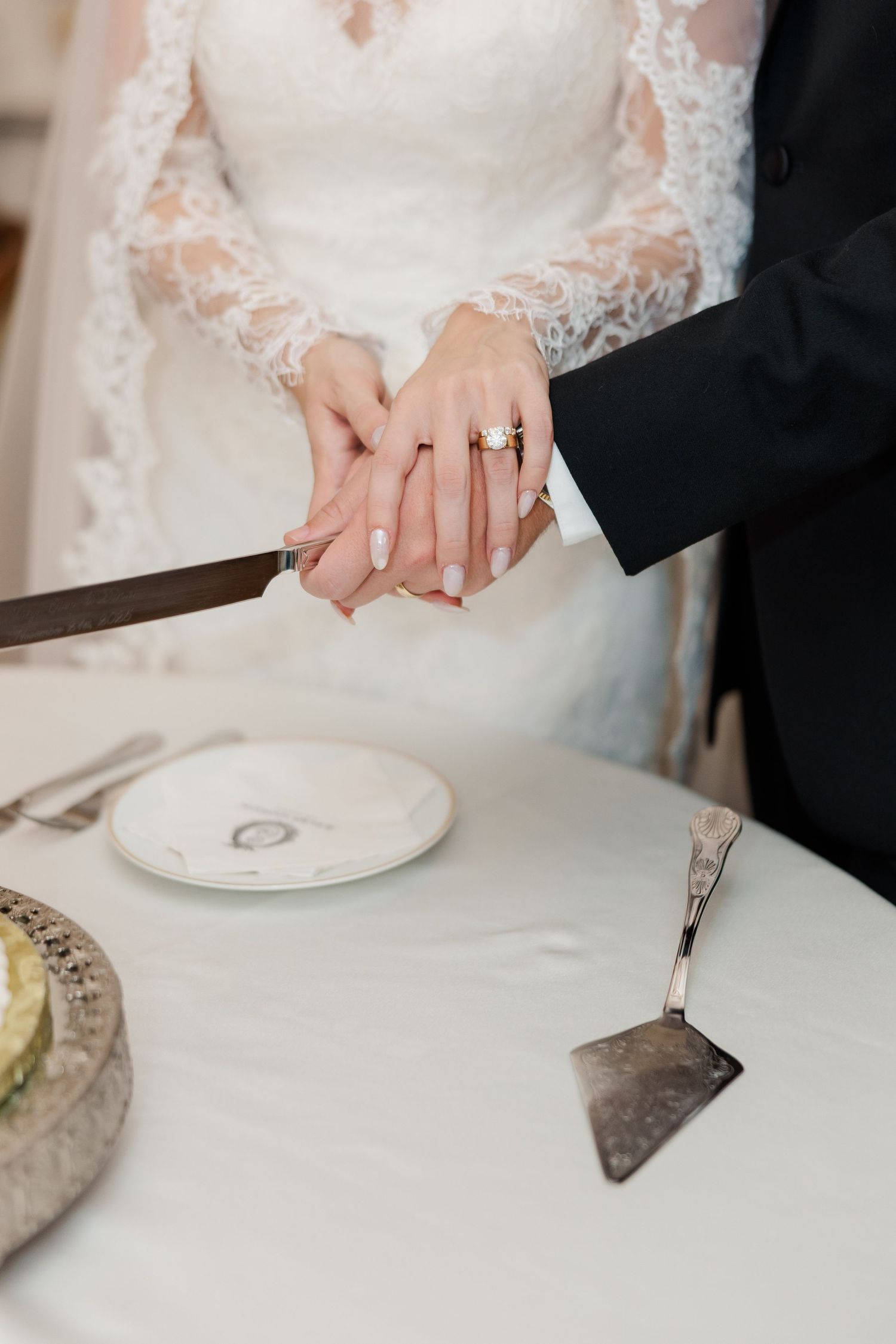 Close-up of hands cutting wedding cake showing engagement ring and lace sleeve detail.
