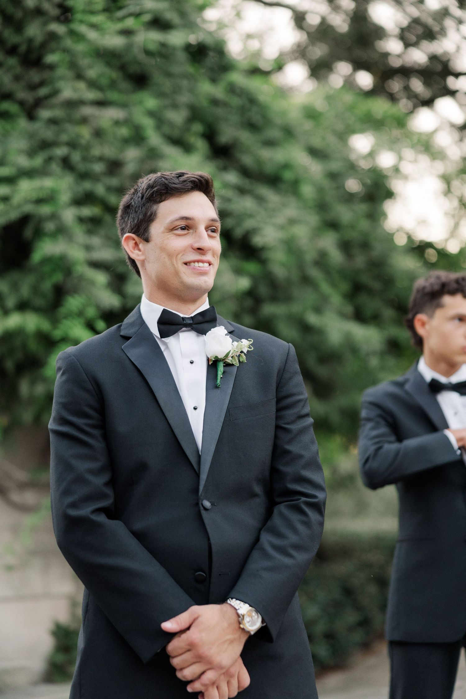 A formal wedding moment outdoors with classic black tuxedos and a boutonniere against lush greenery.