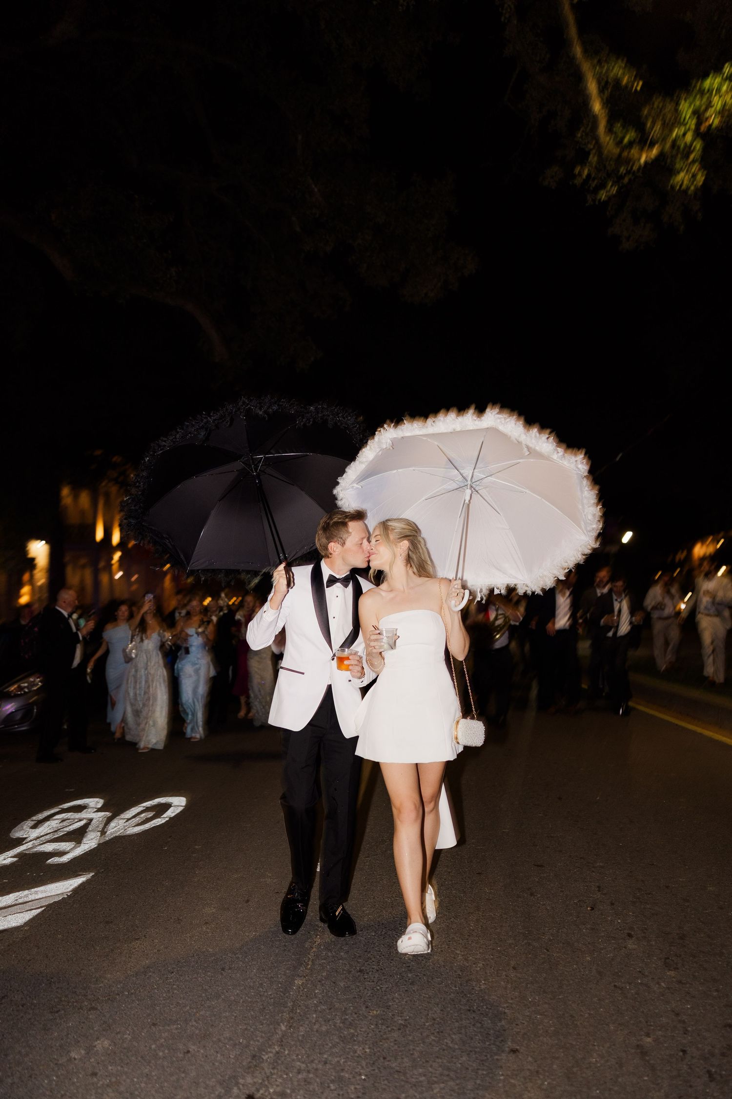 Wedding party members carrying white parasols during an evening celebration outdoors.