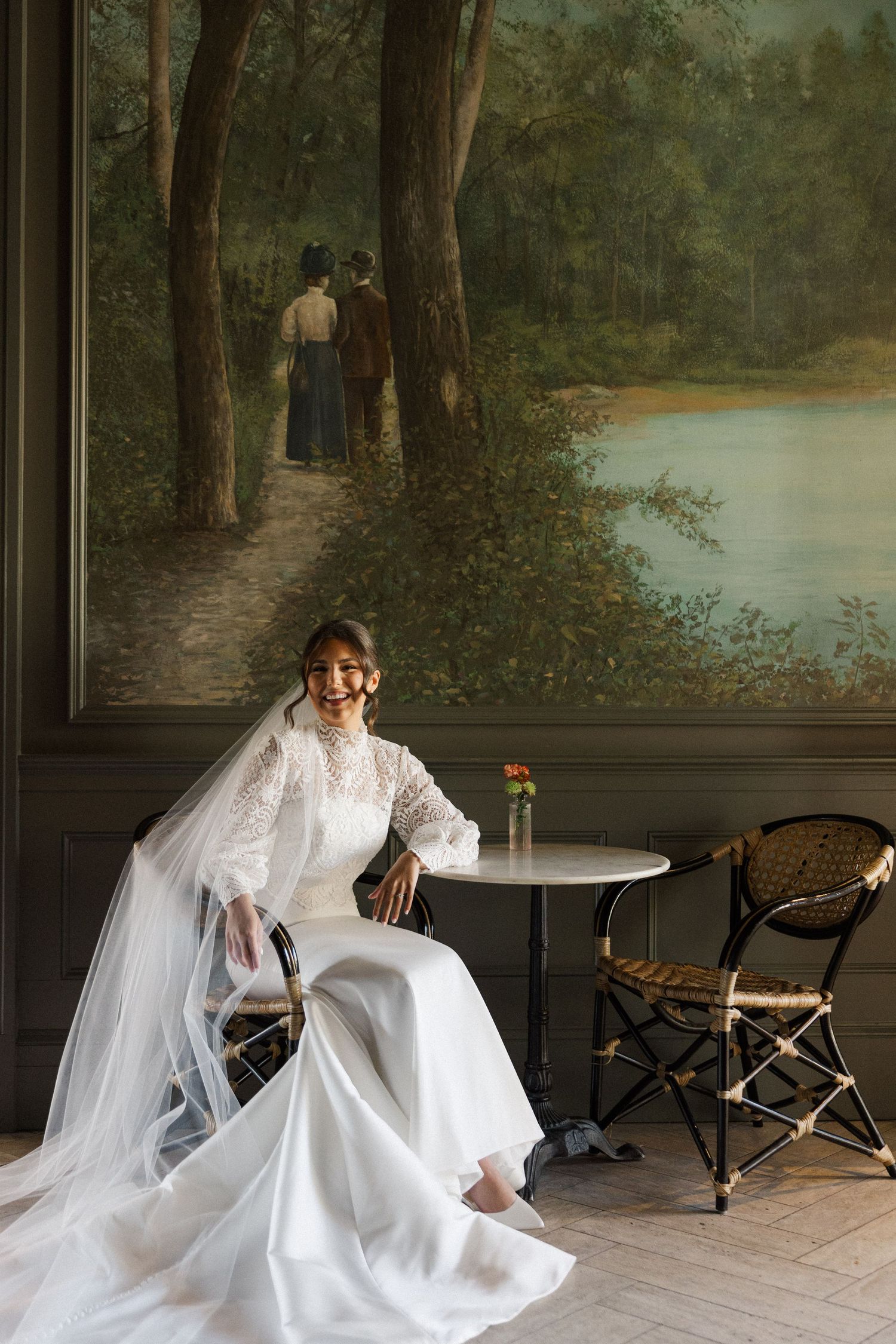 A bride in a flowing white dress and veil poses gracefully on a porch with scenic landscape murals in the background.