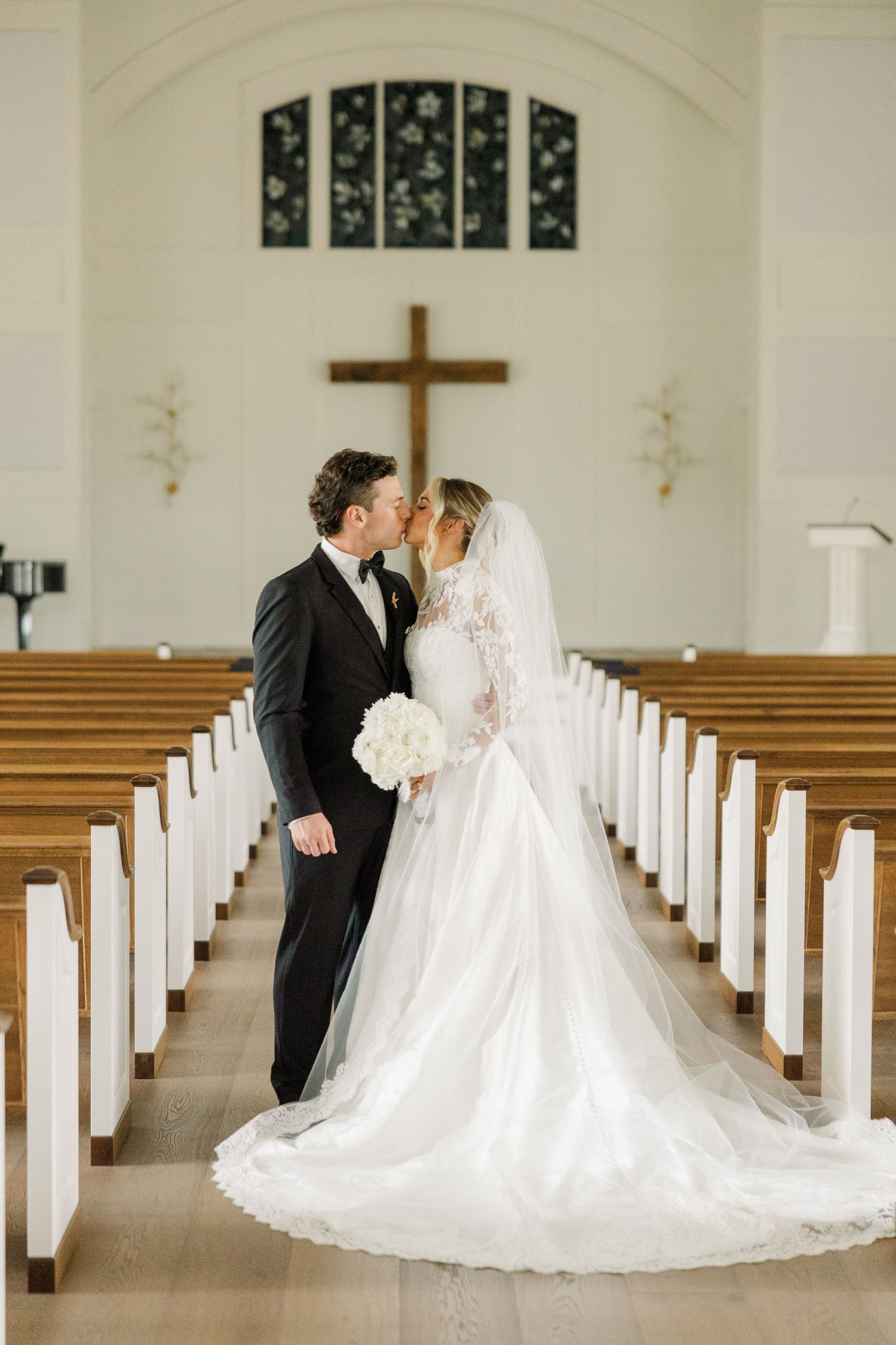 Newlyweds share an intimate moment in a church aisle lined with white pews and a wooden cross in the background.