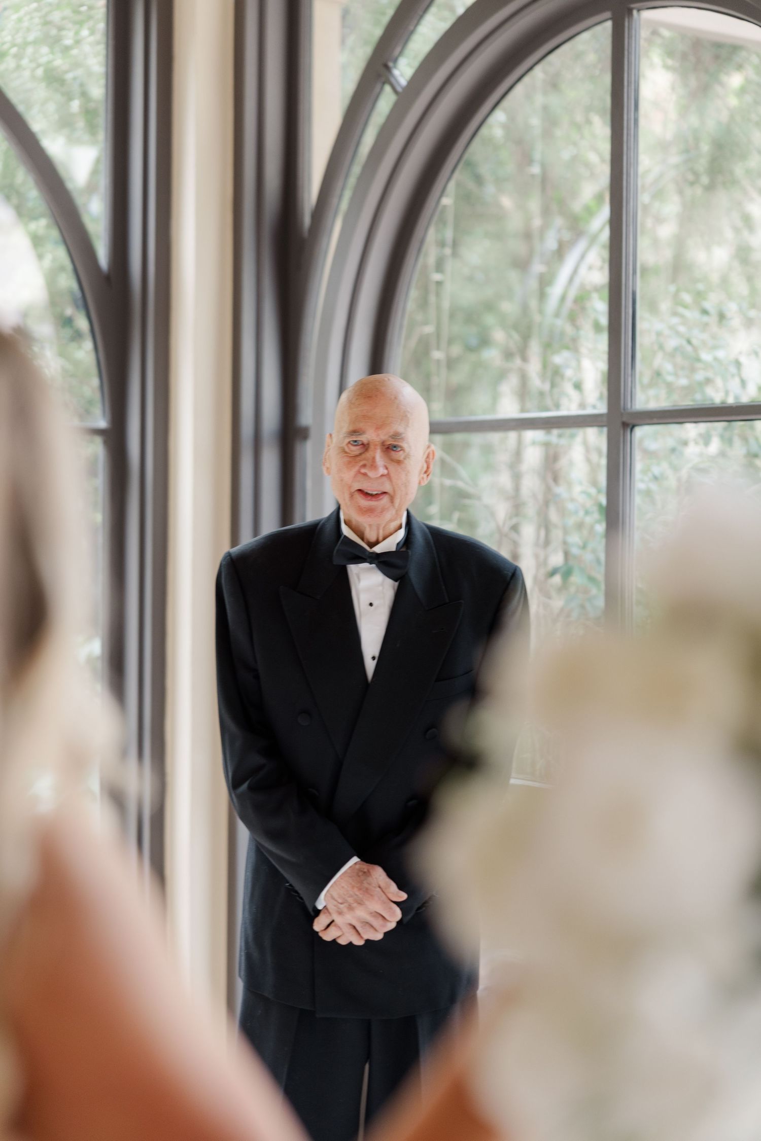 A senior gentleman in a black tuxedo stands in front of an arched window during a formal event.