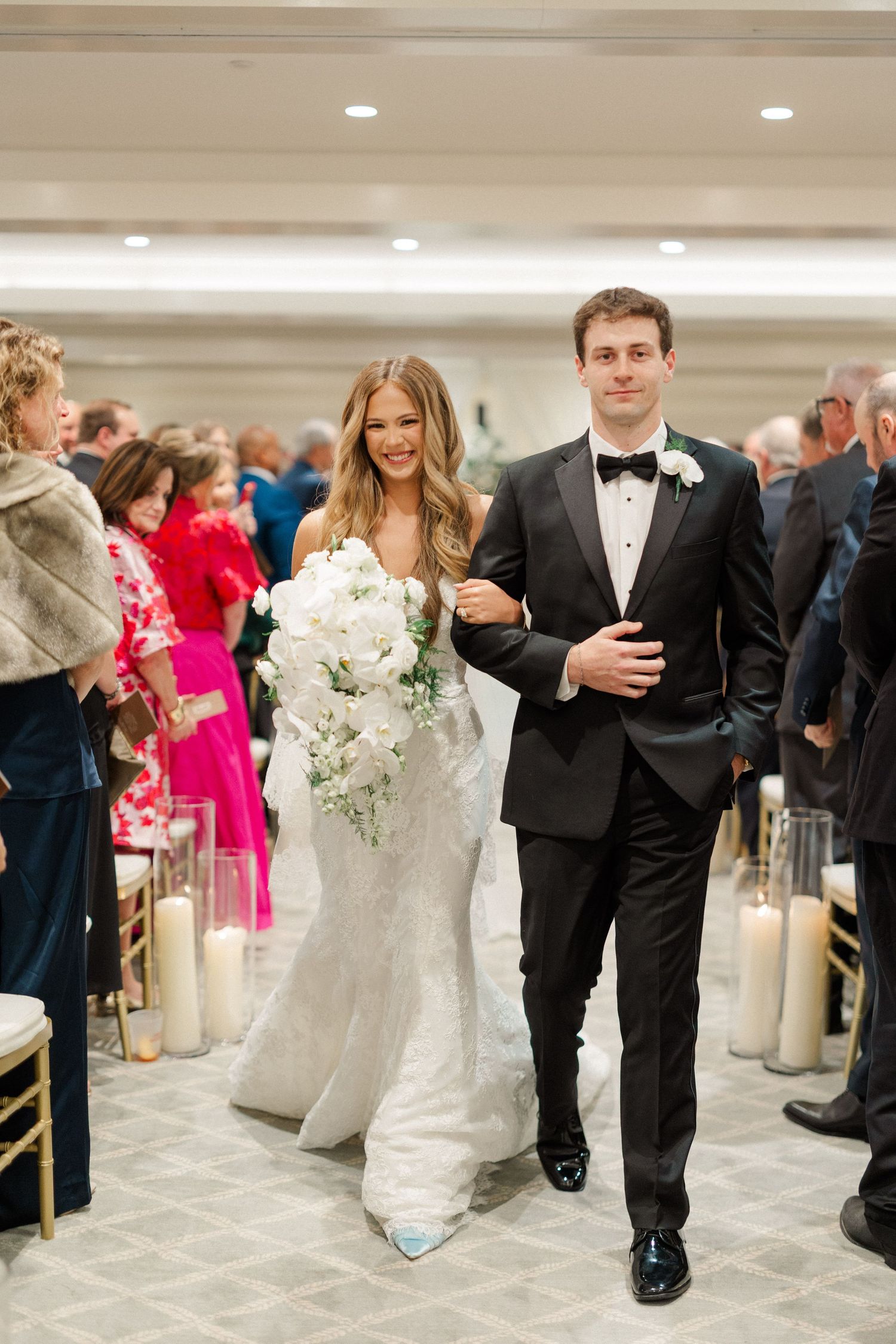 Wedding party members walk down an elegant reception hall aisle lined with guests.