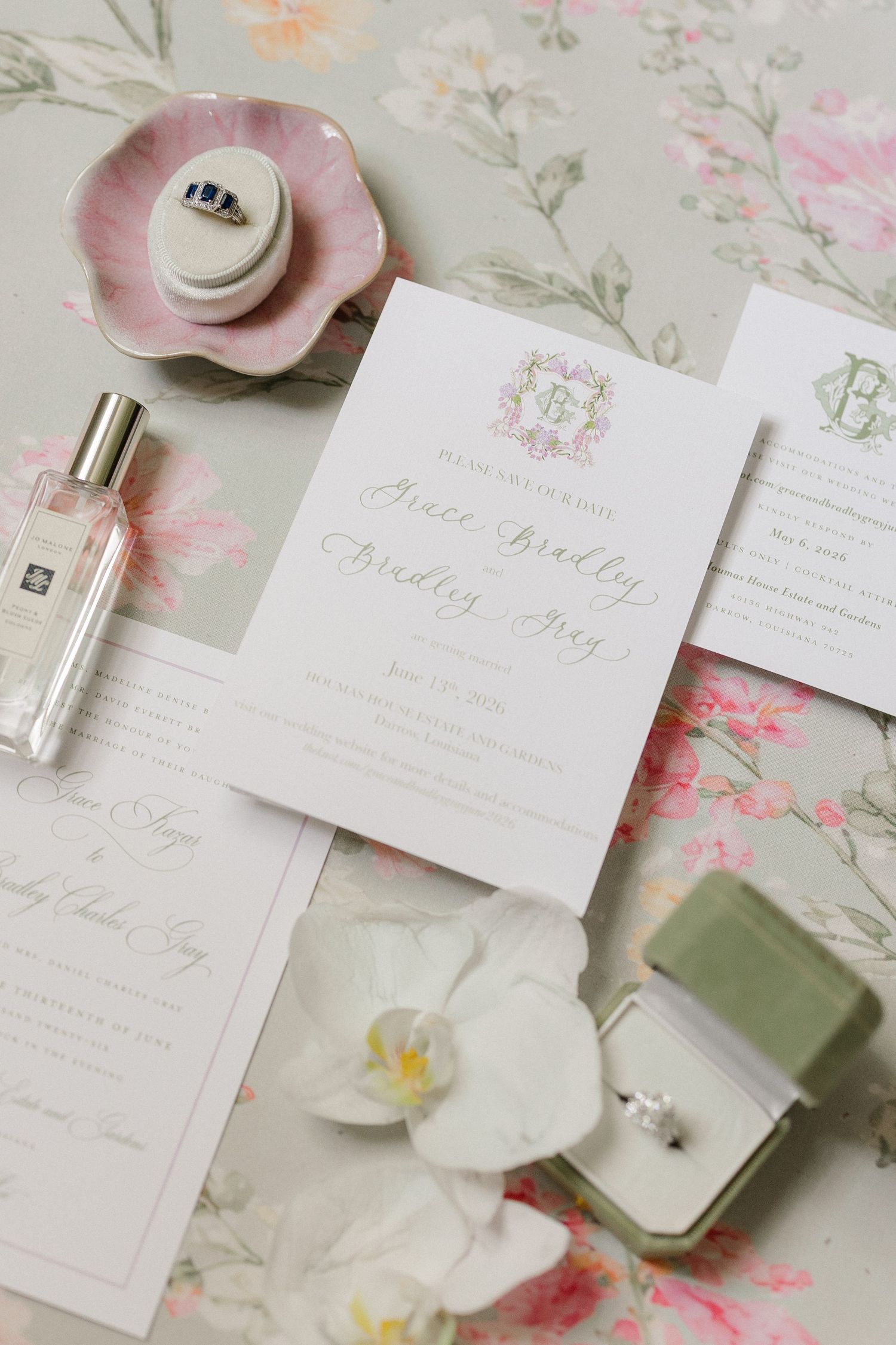 Elegant wedding invitation suite with floral stationery, ring box, perfume bottle, and white orchid on sage green background.