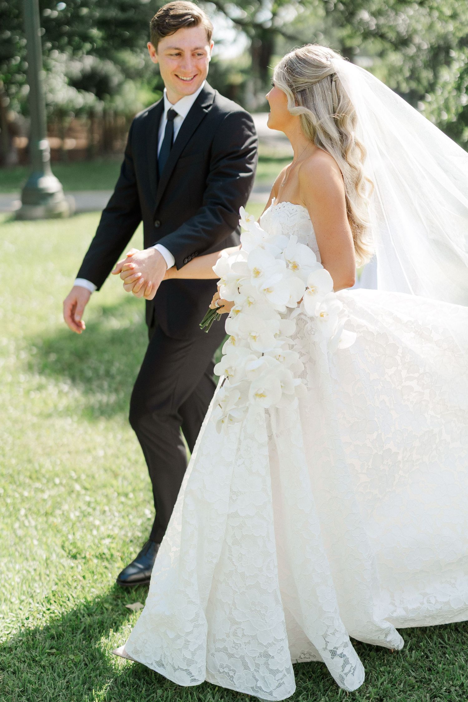 Joyful bride and groom walking hand in hand outdoors, bride wearing lace ballgown and holding white orchid bouquet.