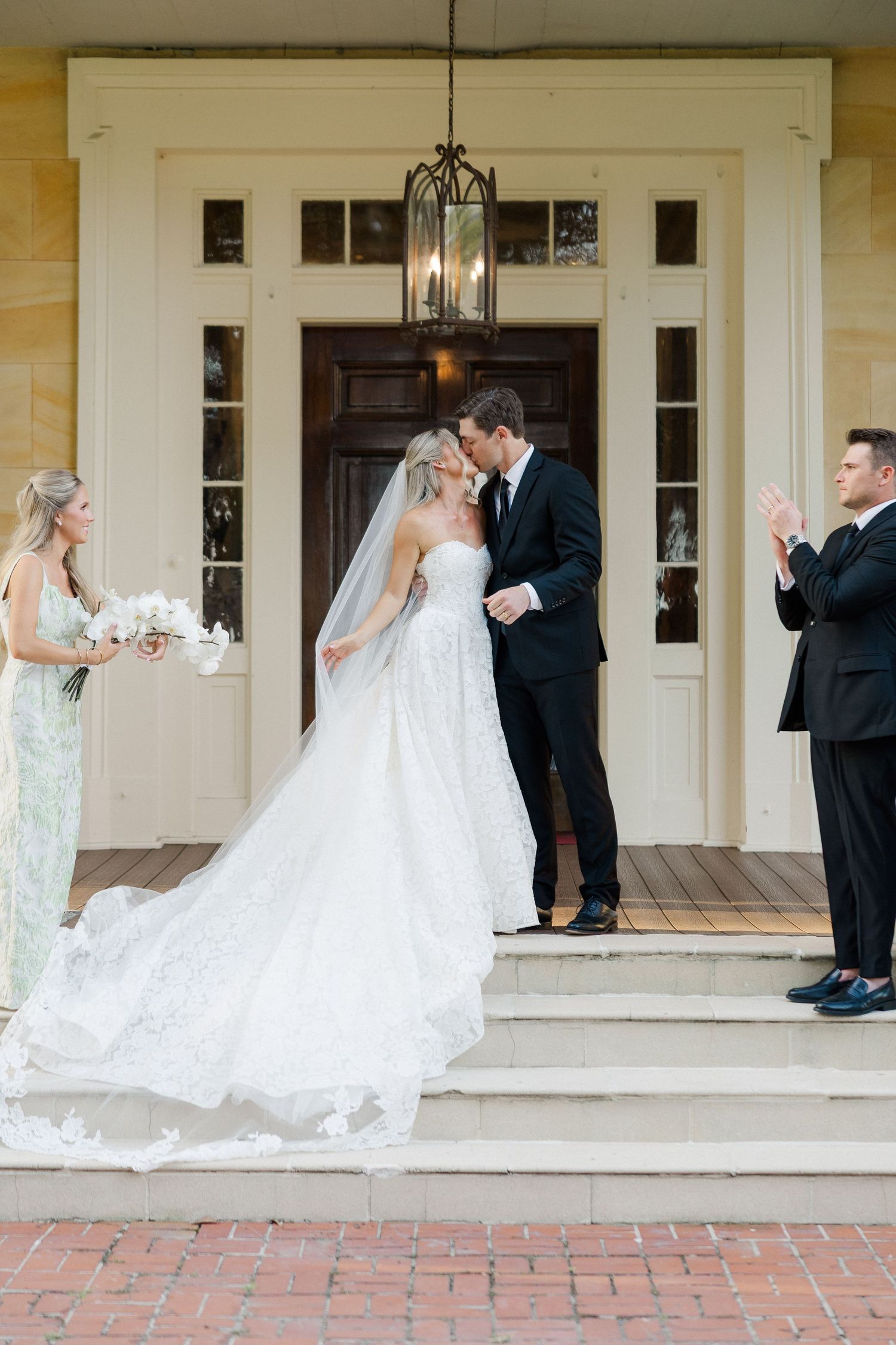 Bride and groom share first kiss on steps of elegant venue as bridesmaid and groomsman applaud beside them.